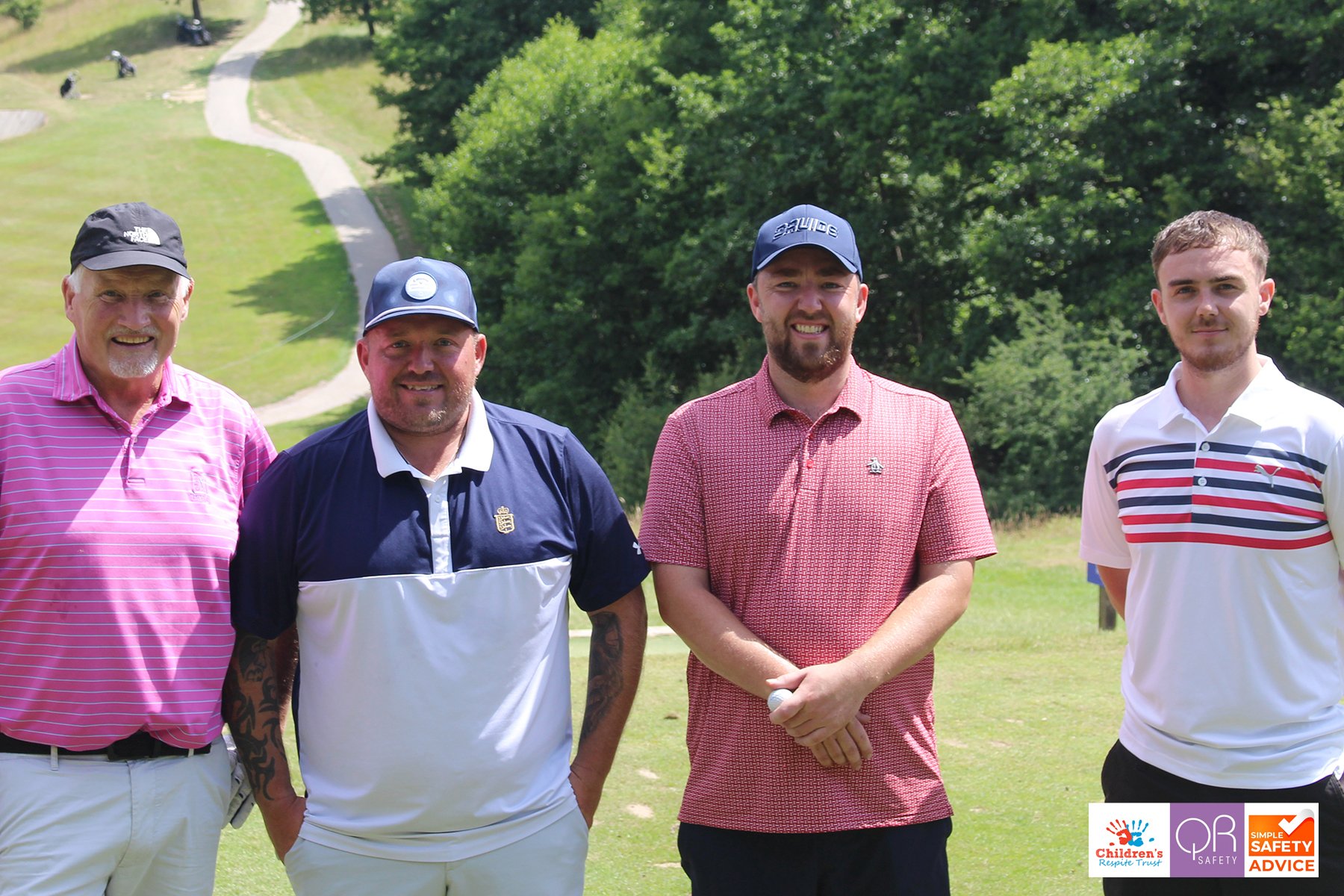 Four men standing on a golf course, smiling, with trees and a winding golf cart path in the background.