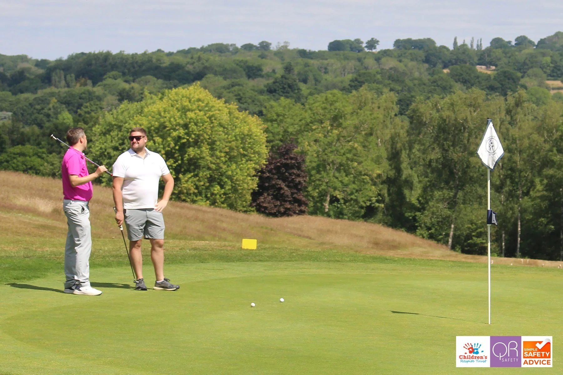 Two men on a golf course discussing, one holding a golf club, with a flag on the green and trees in the background.