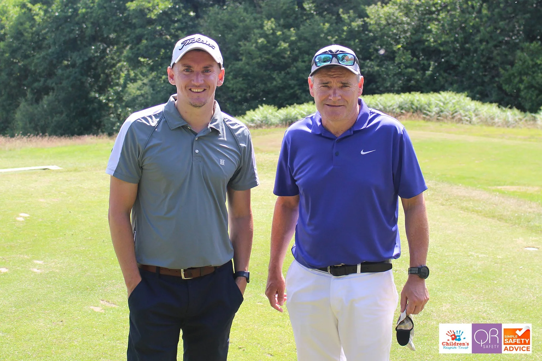 Two men standing on a golf course, smiling at the camera, wearing golf attire and hats, with trees and golf course greenery in the background.