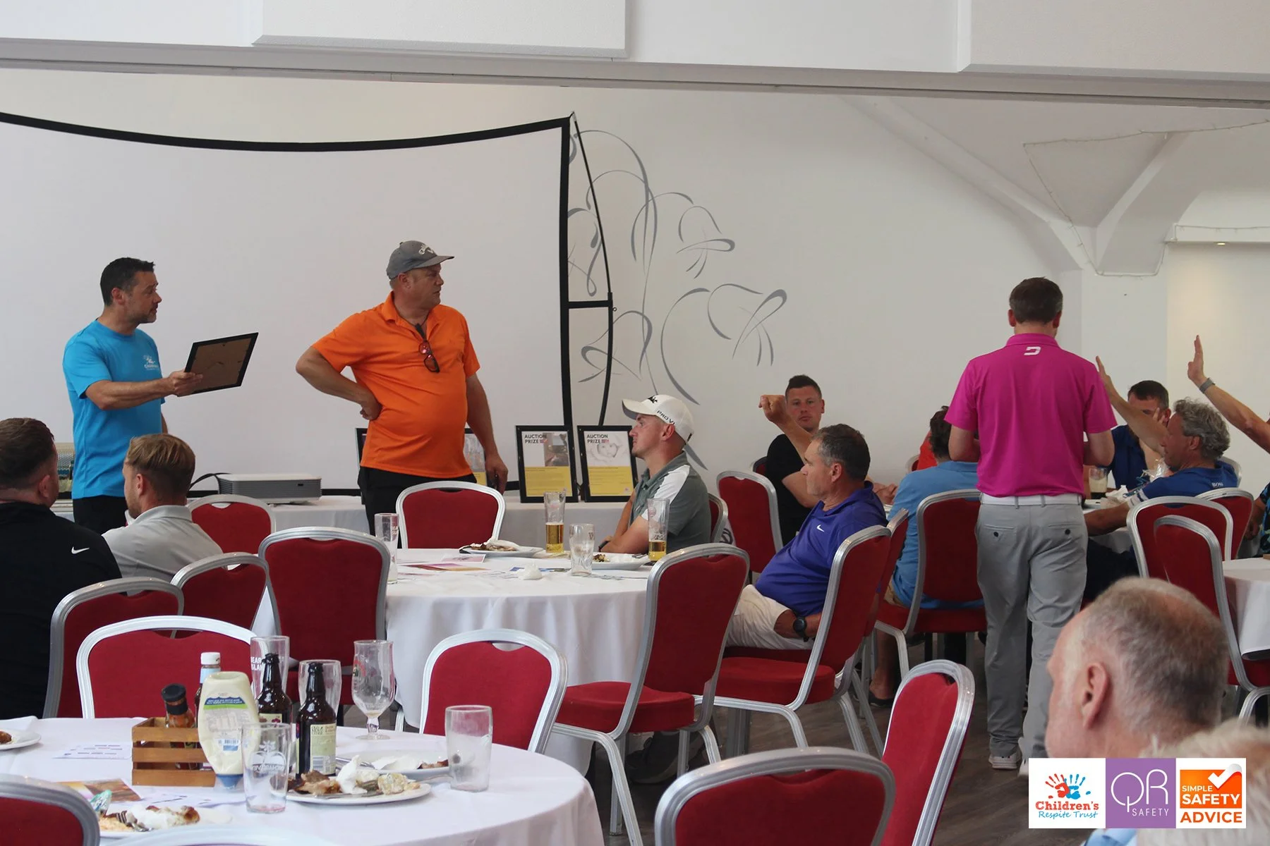 Seated and standing people at a banquet table during an auction event in a large hall, with an auctioneer presenting and people raising hands to bid.