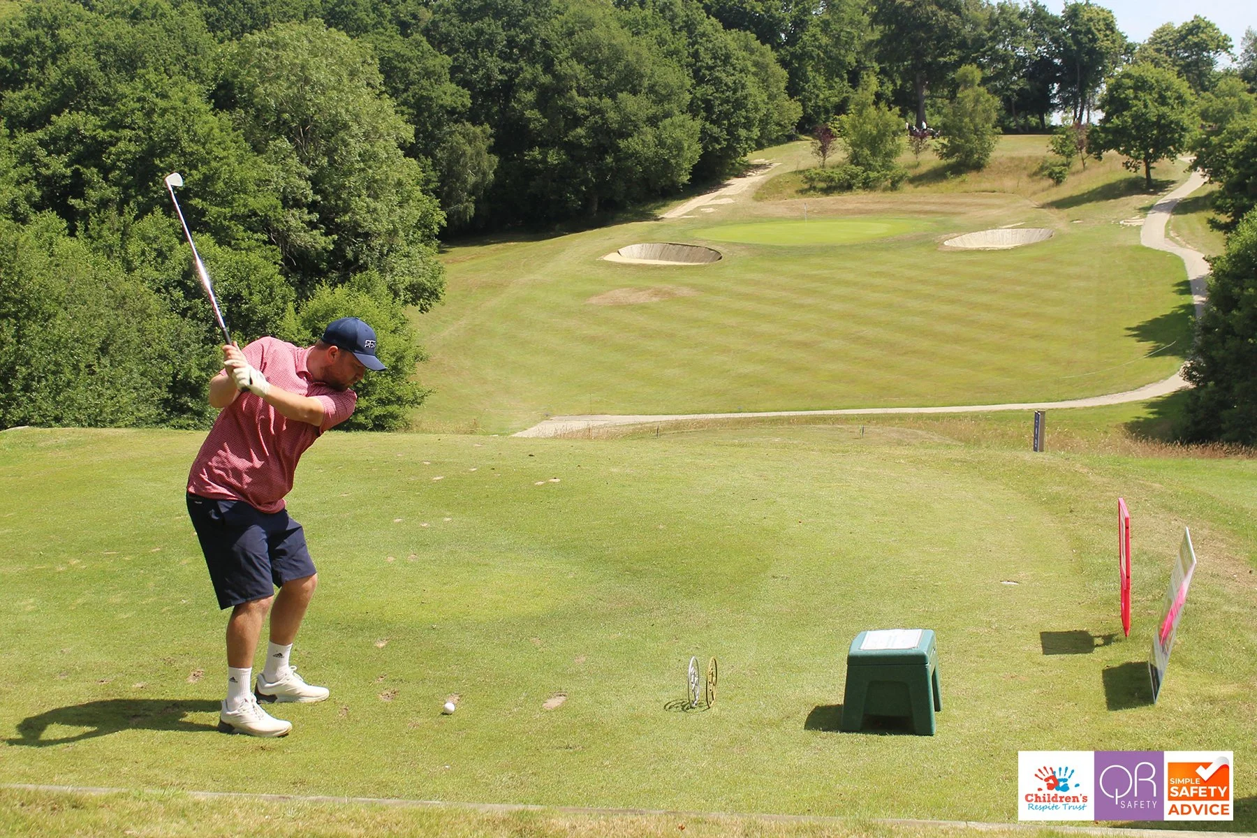 A man wearing a red shirt, black shorts, and a cap prepares to hit a golf ball on the tee. The golf course features multiple sand bunkers and a flag in the distance, surrounded by trees.