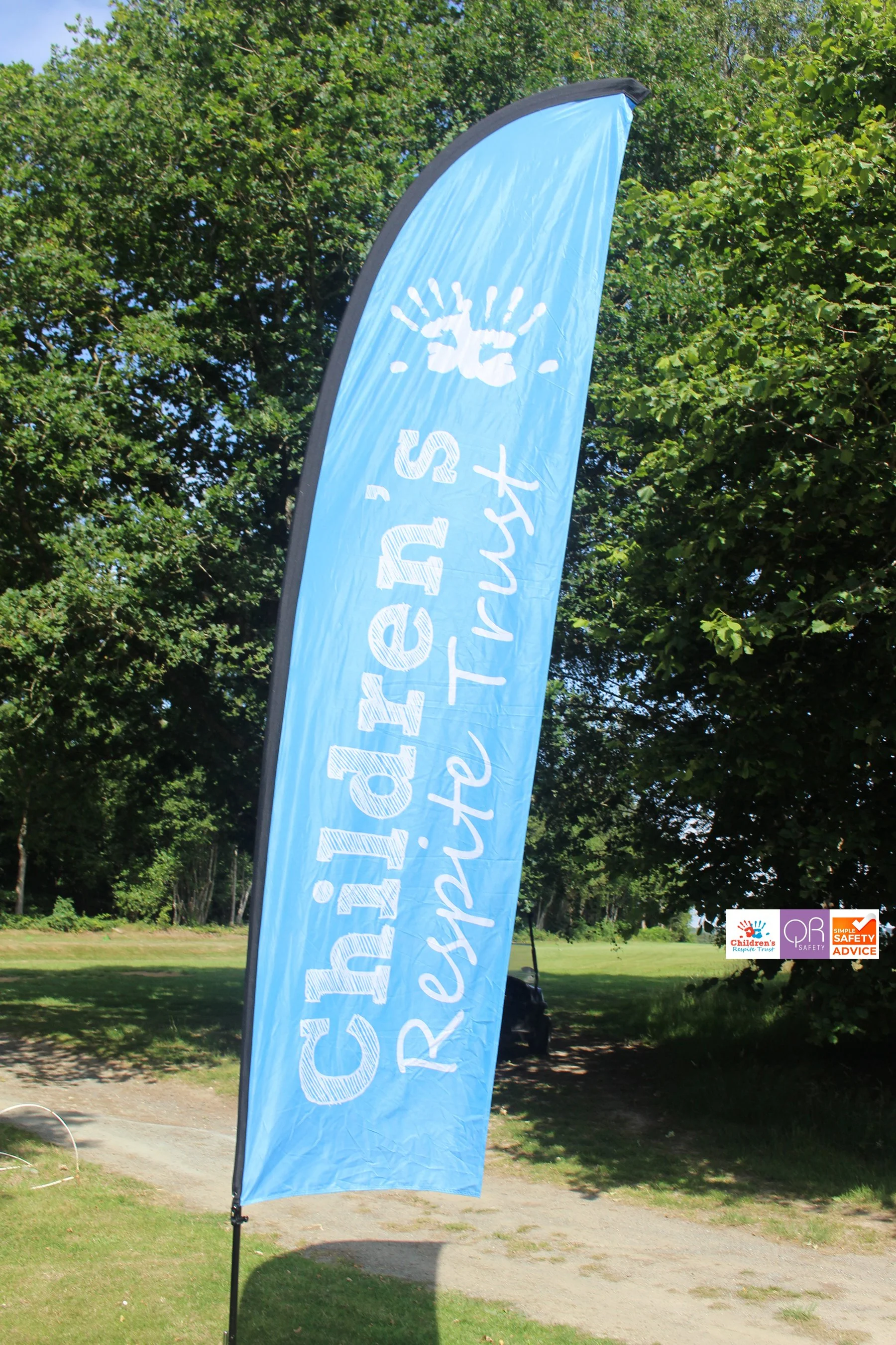 Blue feather flag with white text that reads 'Children's Respite Trust' and a sun logo, standing outdoors on grass with trees in the background.