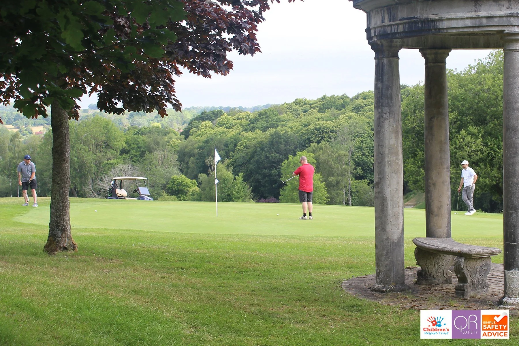 Three men playing golf on a lush green golf course with trees in the background. One man in a red shirt is about to hit the golf ball, while the others observe. A golf cart is parked near the hole with a flag, and a stone bench with columns is visibl