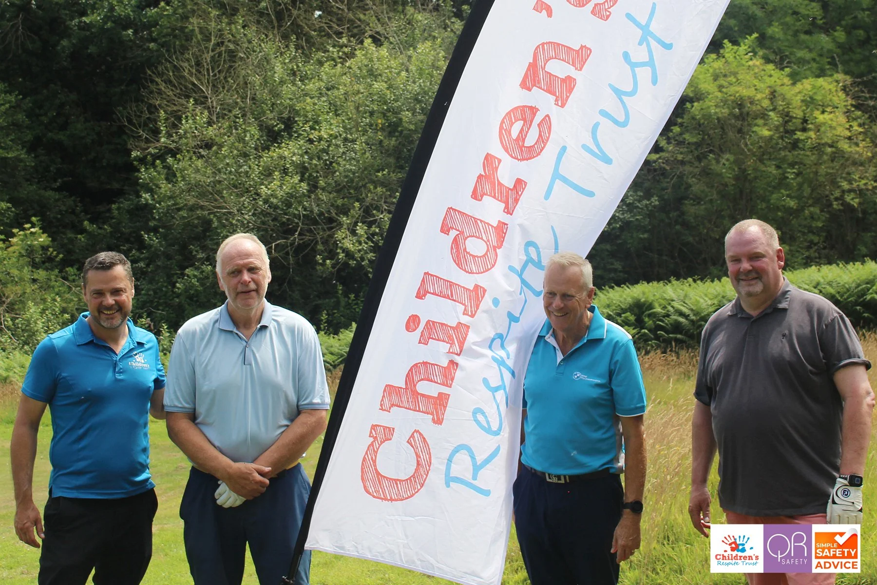 Four men standing outdoors on a grassy area behind a large banner that reads "Children's Response Trust" with logos of children's safety and safety advice organizations at the bottom right corner, with a background of green trees and bushes.