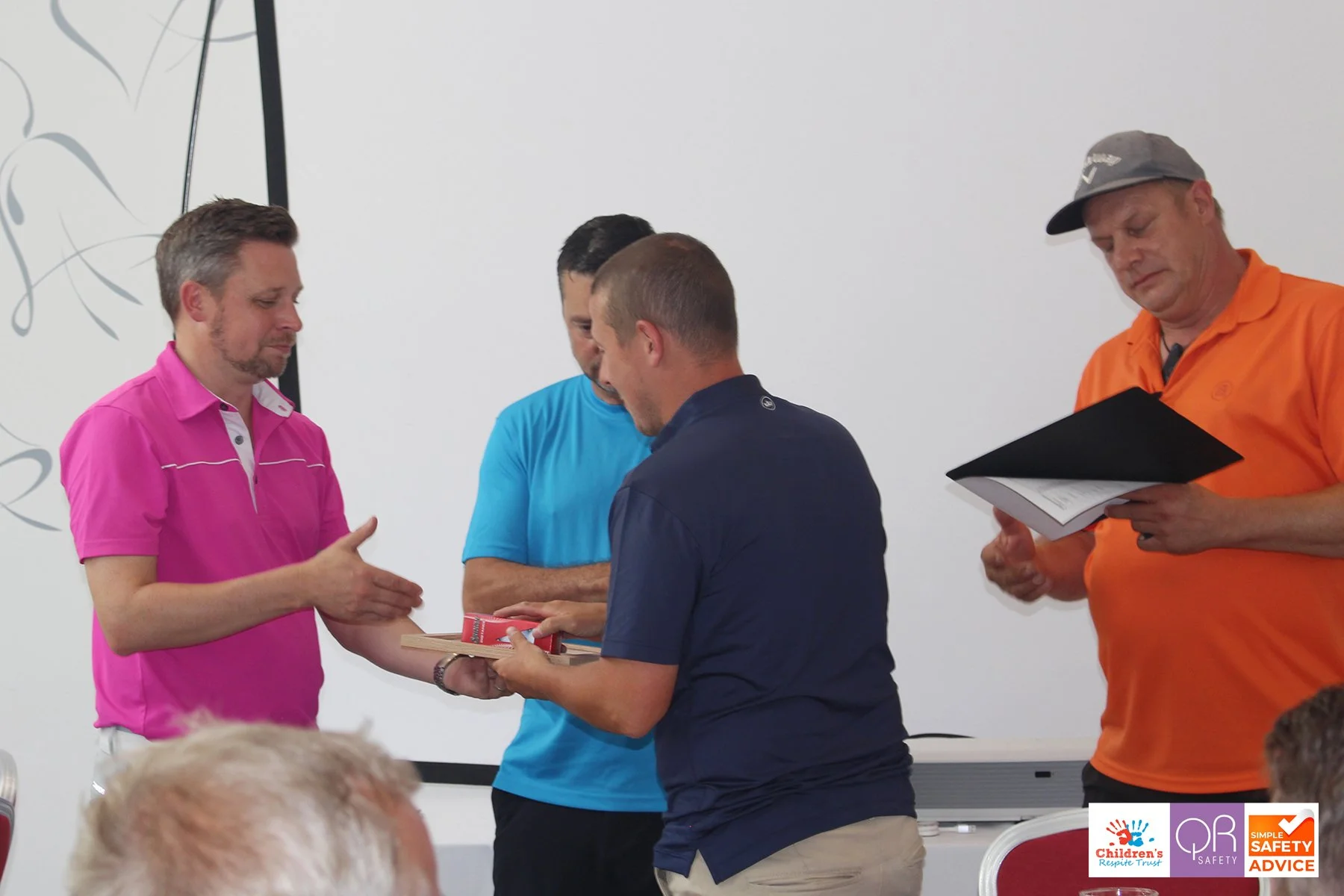 A man in a pink shirt is shaking hands with another man in a blue shirt, while two other men watch, one holding a certificate or award. The setting appears to be an indoor event or ceremony.