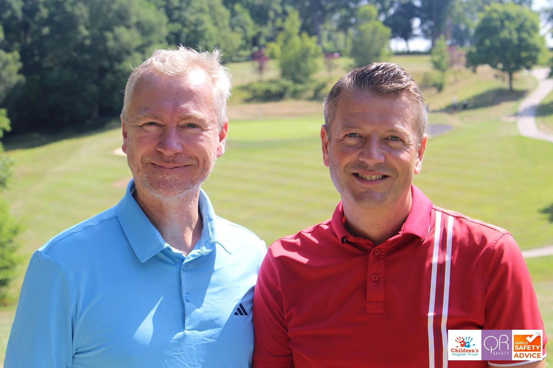 Two smiling men outdoors on a sunny day, standing on a grassy area with trees and a path in the background, wearing collared shirts, one blue and one red.