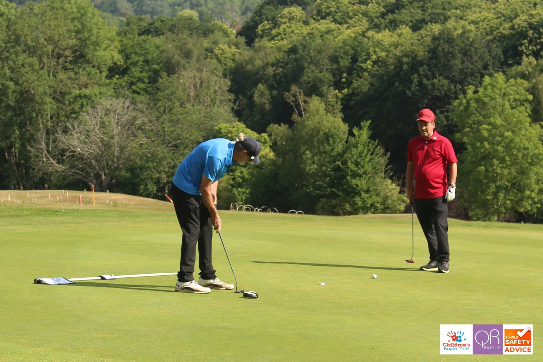 Two men playing golf on a lush green golf course with trees in the background. One man is preparing to putt, while the other stands nearby observing. The man putting is wearing a blue shirt, black pants, and a black cap. The other man is wearing a re