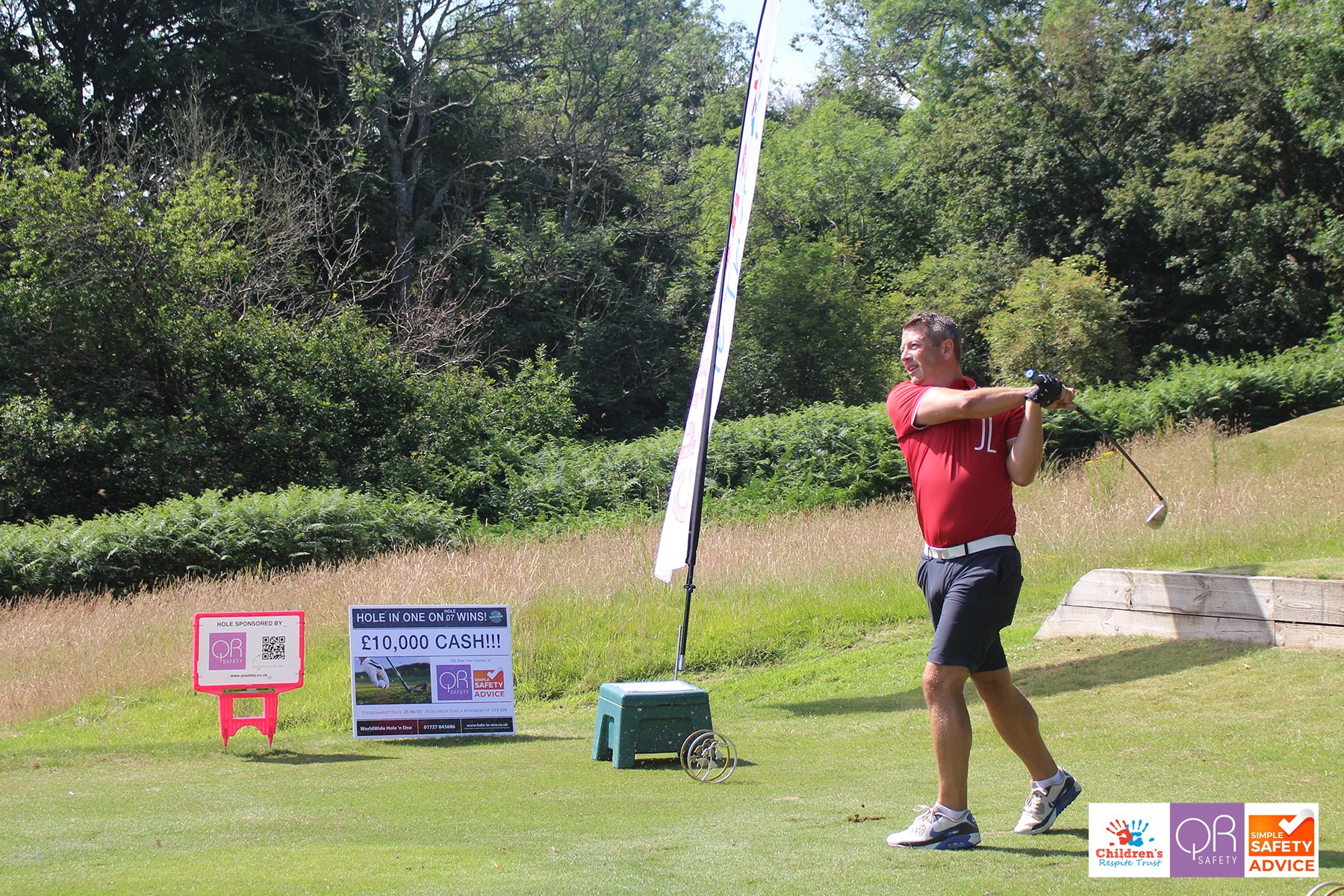 A man in a red shirt and black shorts swinging a golf club on a golf course, with signs and a flag nearby, surrounded by greenery.