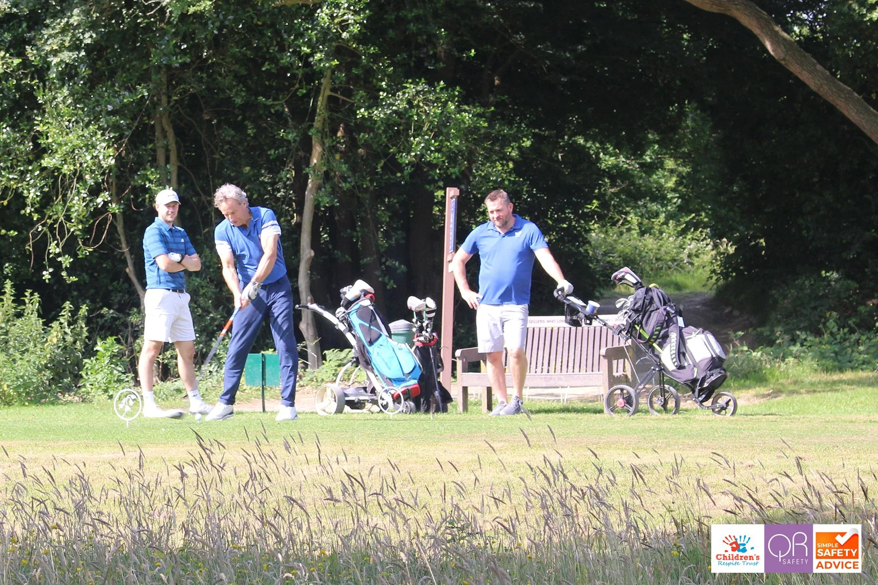 Three men playing golf on a sunny fairway, with golf clubs and carts nearby, trees in the background, and safety and children's trust logos in the corner.