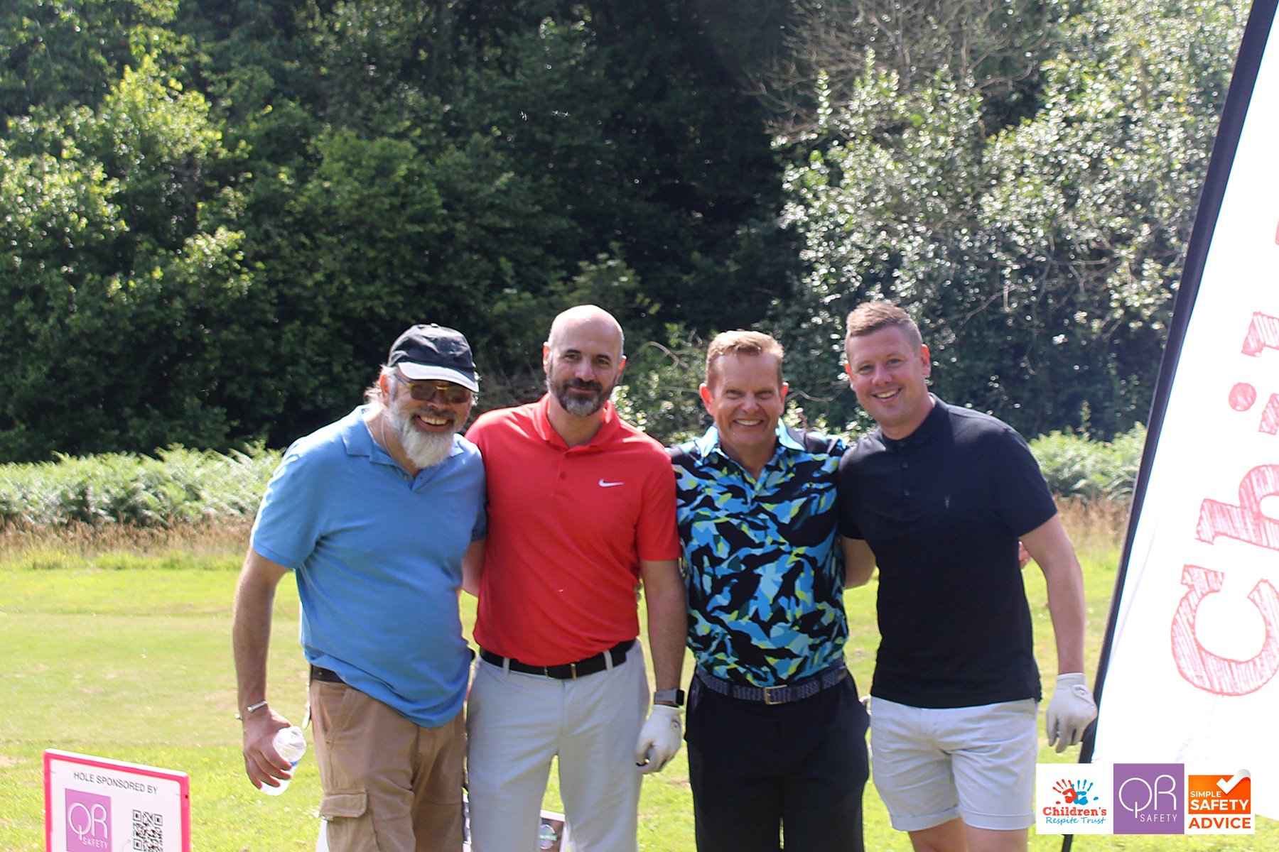 Four men smiling at a golf course, standing close together, dressed in golf attire with trees in the background.