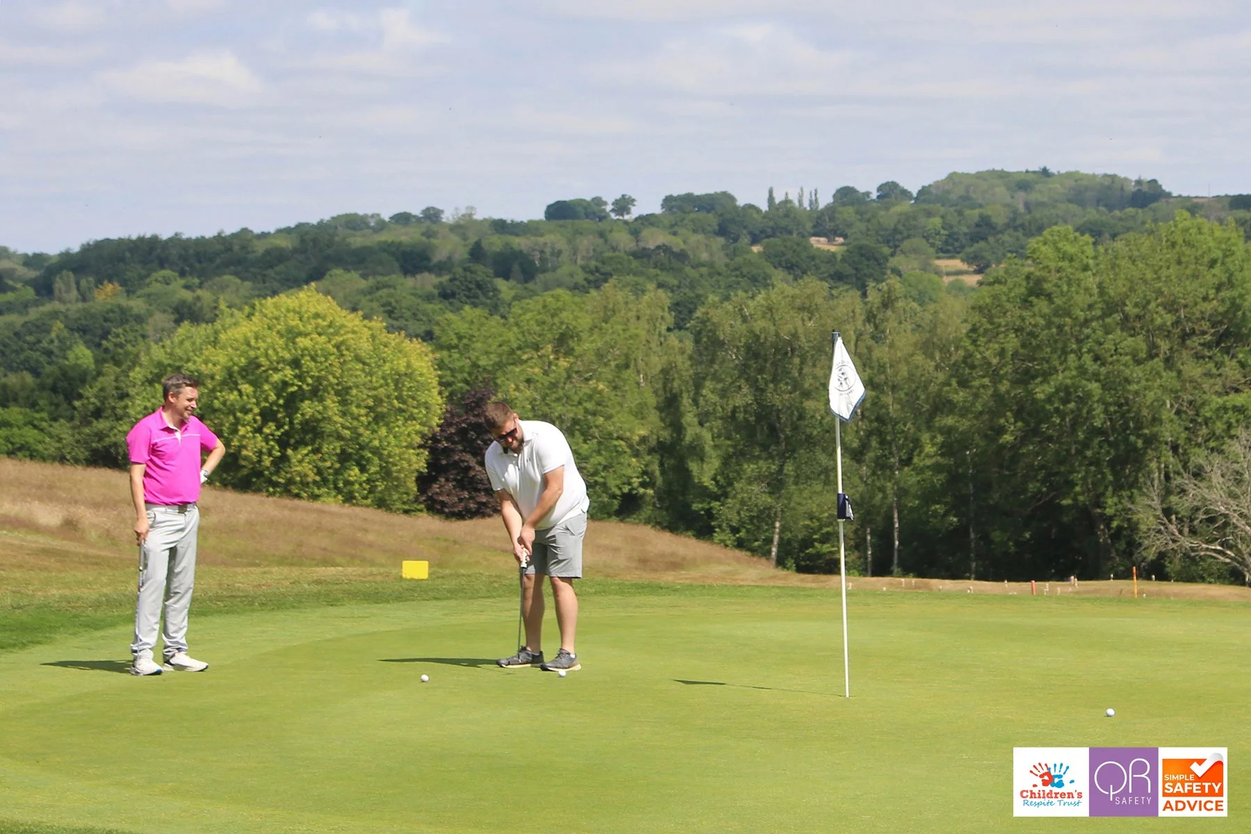 Two men playing golf on a lush green golf course with trees, rolling hills, and a partly cloudy sky in the background. One man stands observing as the other prepares to putt near a hole marked by a flagstick.