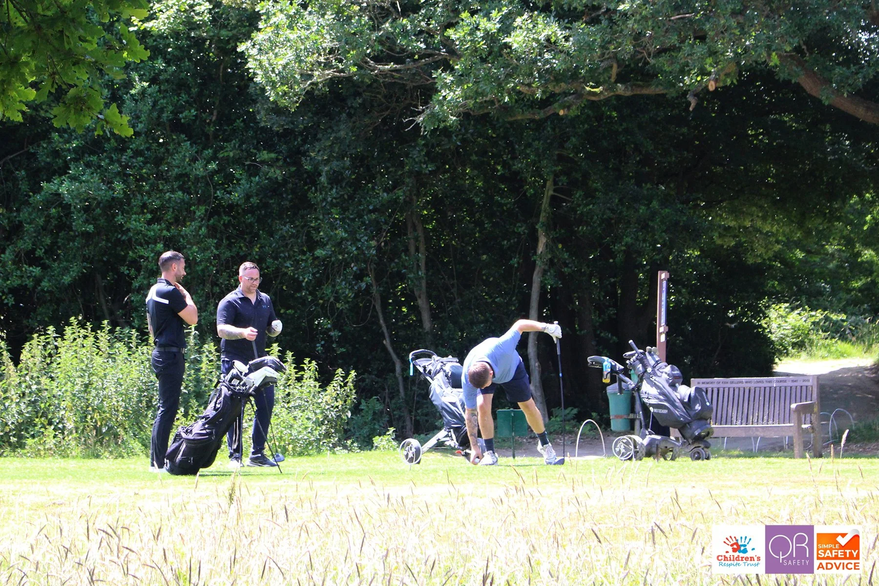 Two men on a golf course with golf bags and a golf cart, one is bending over to play golf, while the other two are standing nearby watching, with a tree line in the background.