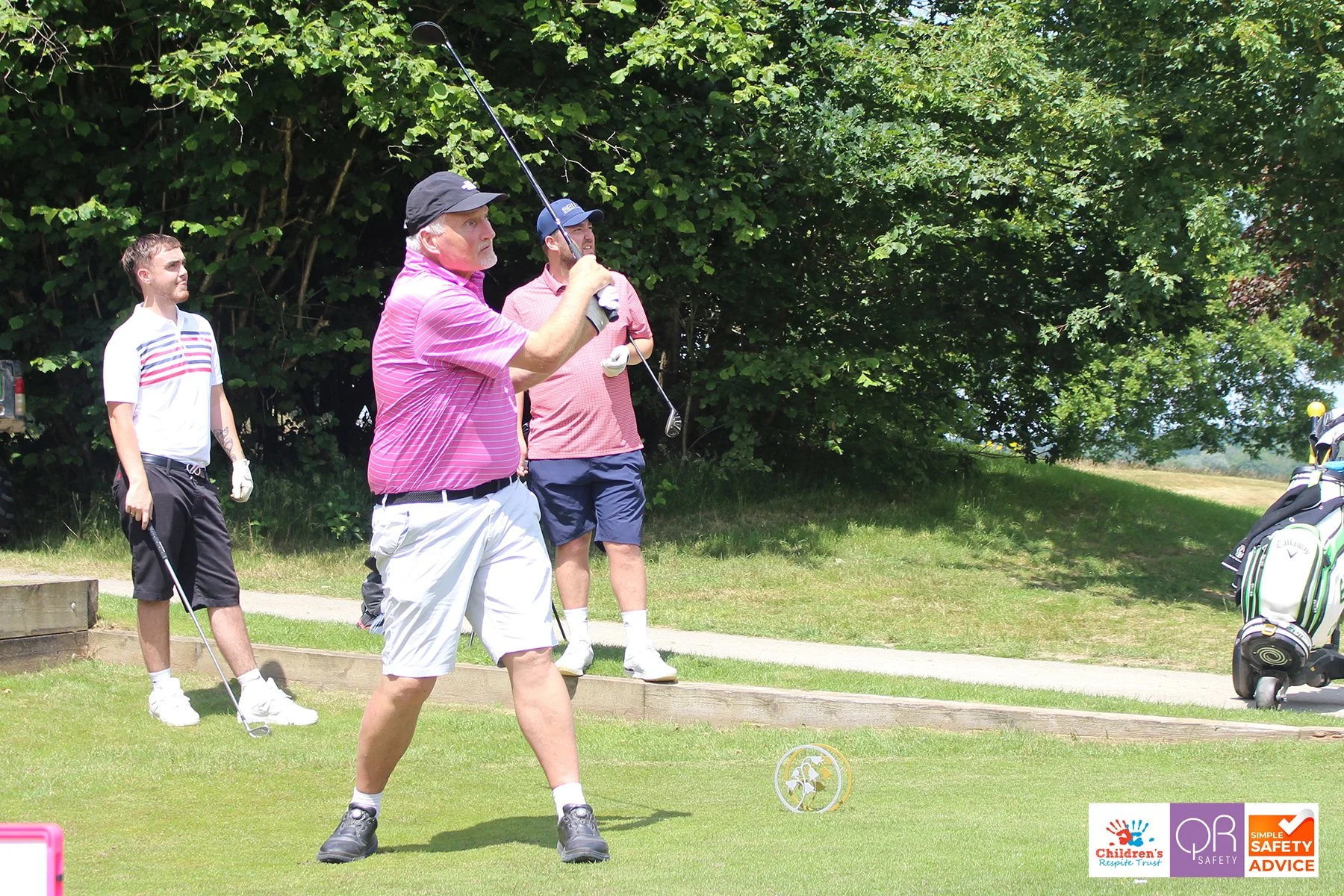 Three men on a golf course; one is in the process of swinging a golf club while the others watch. There are trees and a golf bag in the background.