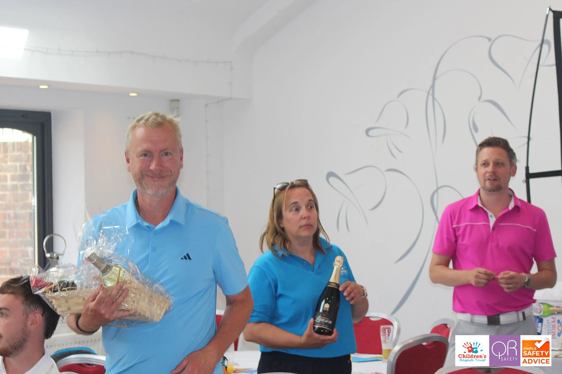 Three people standing indoors, holding gifts. The man on the left holds a basket with a bottle, the woman in the middle holds a large bottle of champagne, and the man on the right stands in front of a white wall with minimal decoration.