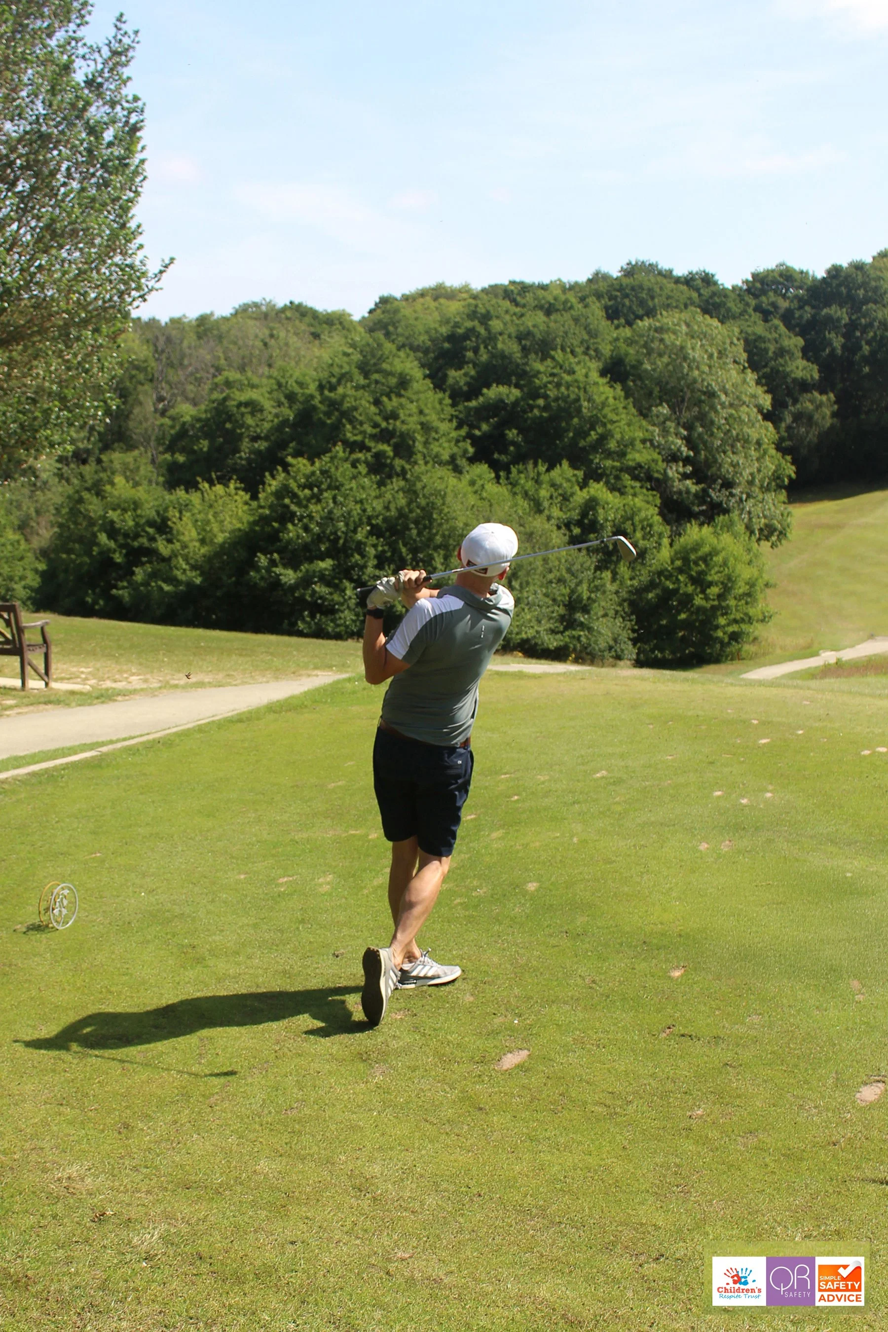 A man in a gray T-shirt, navy shorts, and a white cap swinging a golf club on a lush green golf course surrounded by trees and clear blue sky.
