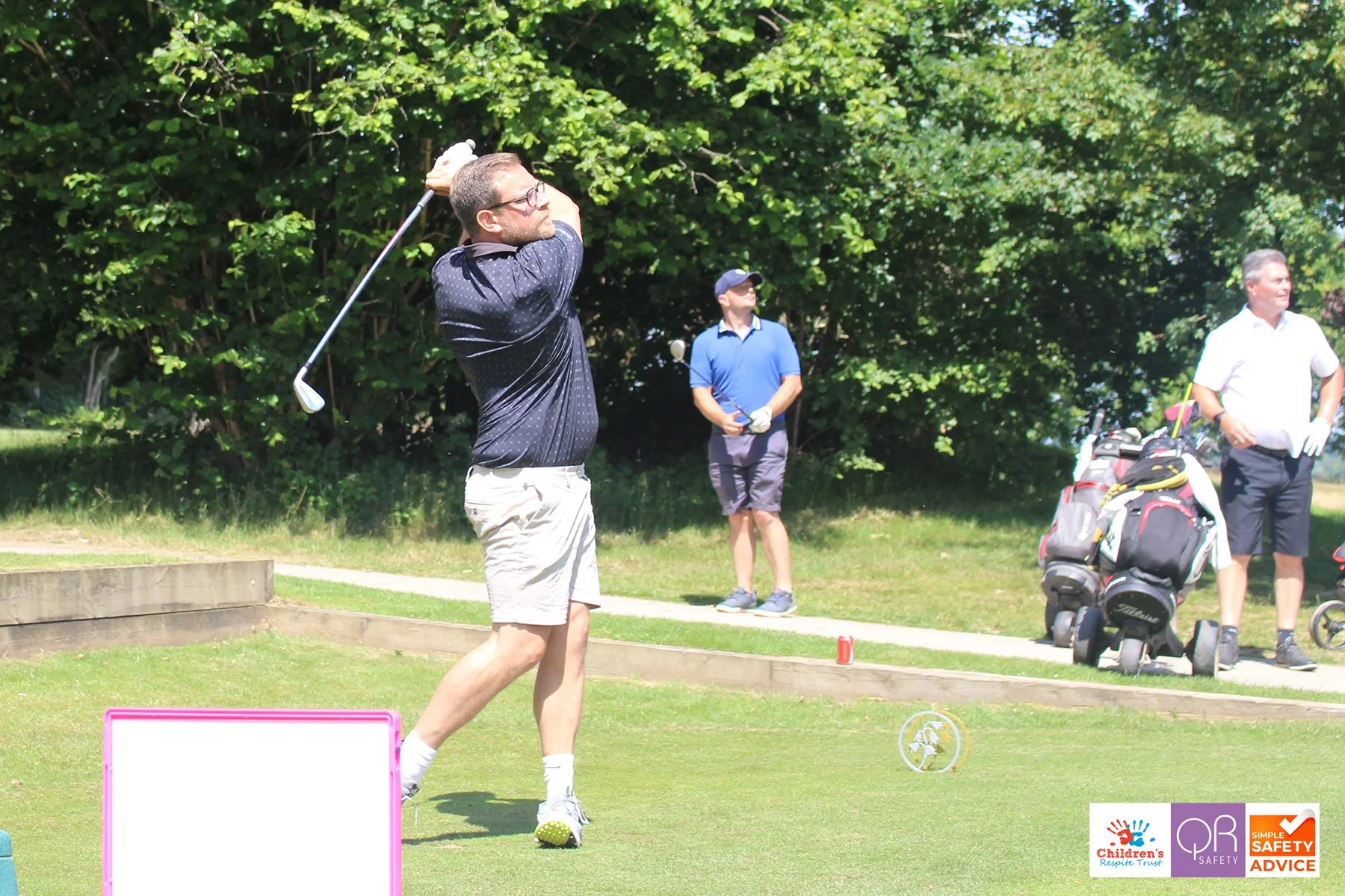 Man playing golf on a grassy course, swinging a golf club, with other men and golf bags in the background, on a sunny day with trees.
