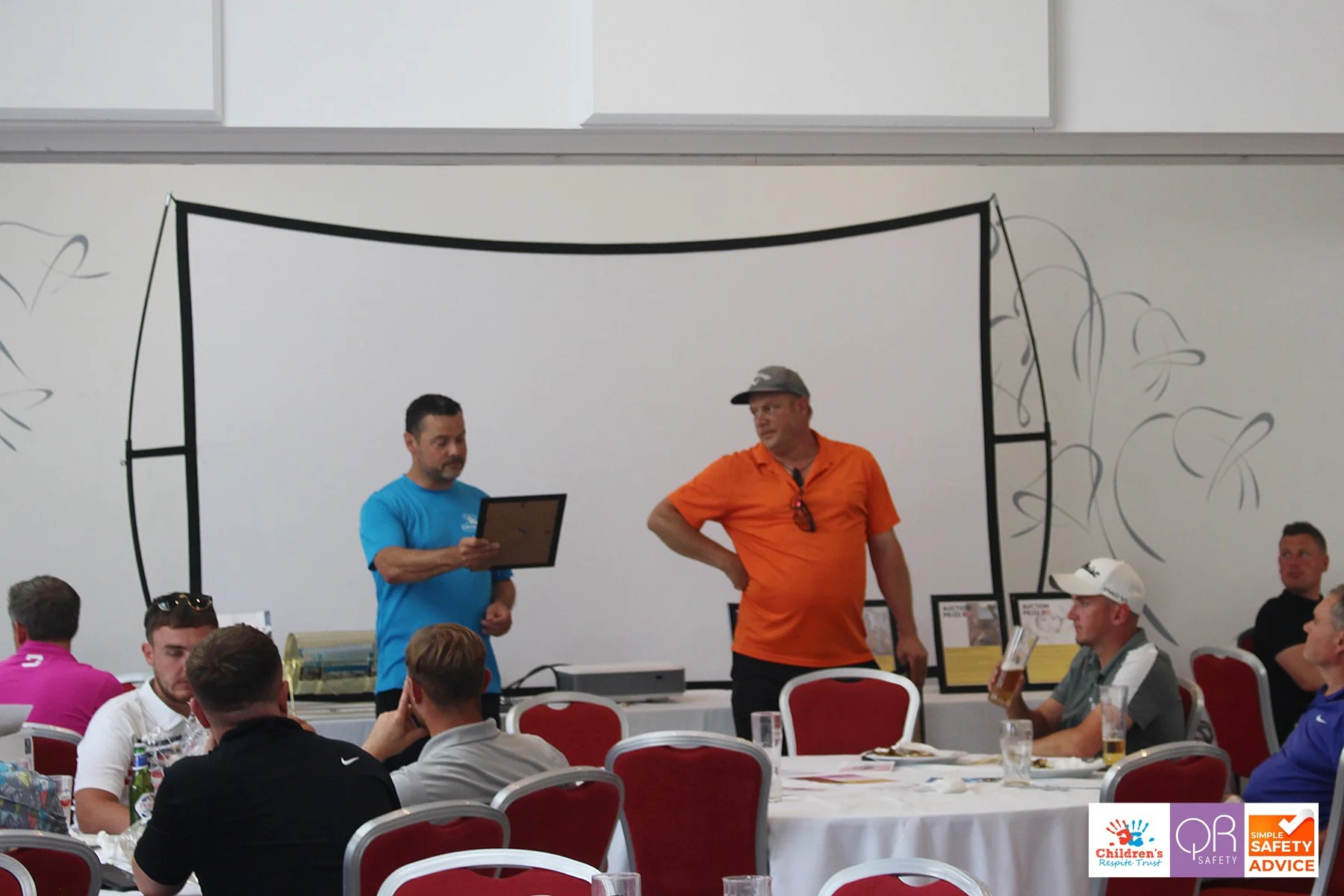 People sitting at round tables with white tablecloths in a banquet hall, two men standing near the wall, one holding a framed certificate, the other wearing an orange shirt and cap. Some attendees are drinking beer and eating, with logos of Children'