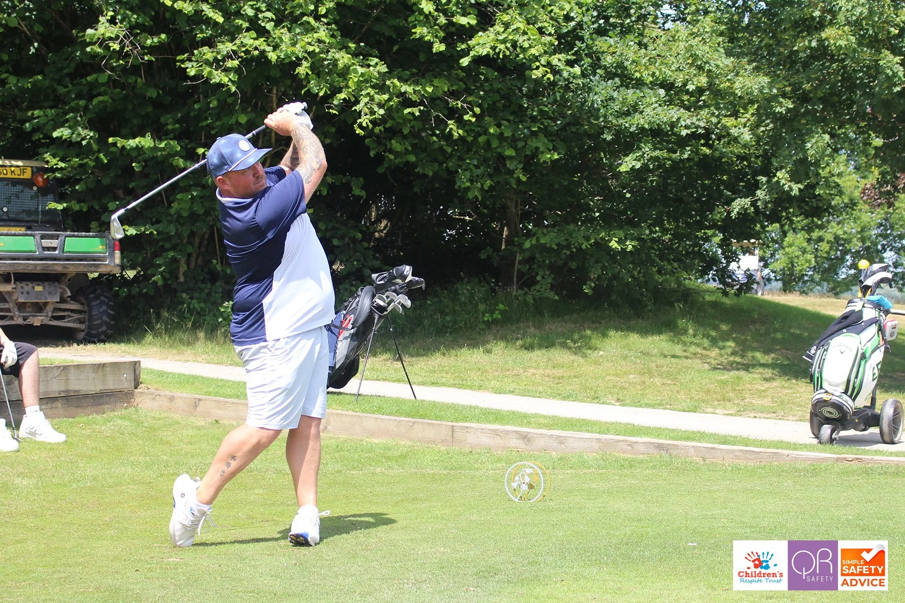 A man golfing on a golf course, swinging a golf club. He is wearing a blue and white shirt, white shorts, a blue cap, and white shoes. There are trees in the background and a golf bag with clubs on a pushcart to his right. Logos for Children's Resort