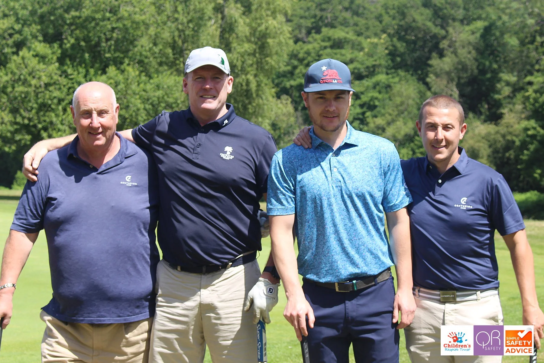 Four men standing arm-in-arm on a golf course, dressed in golf attire, with trees in the background.