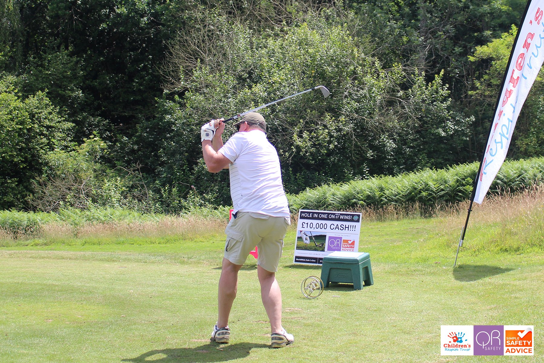 A man in shorts and a white shirt is swinging a golf club on a golf course. There is a sign advertising a cash prize of 10,000 pounds and a flag on the right side with logos from children's charity and safety organizations. The background features gr