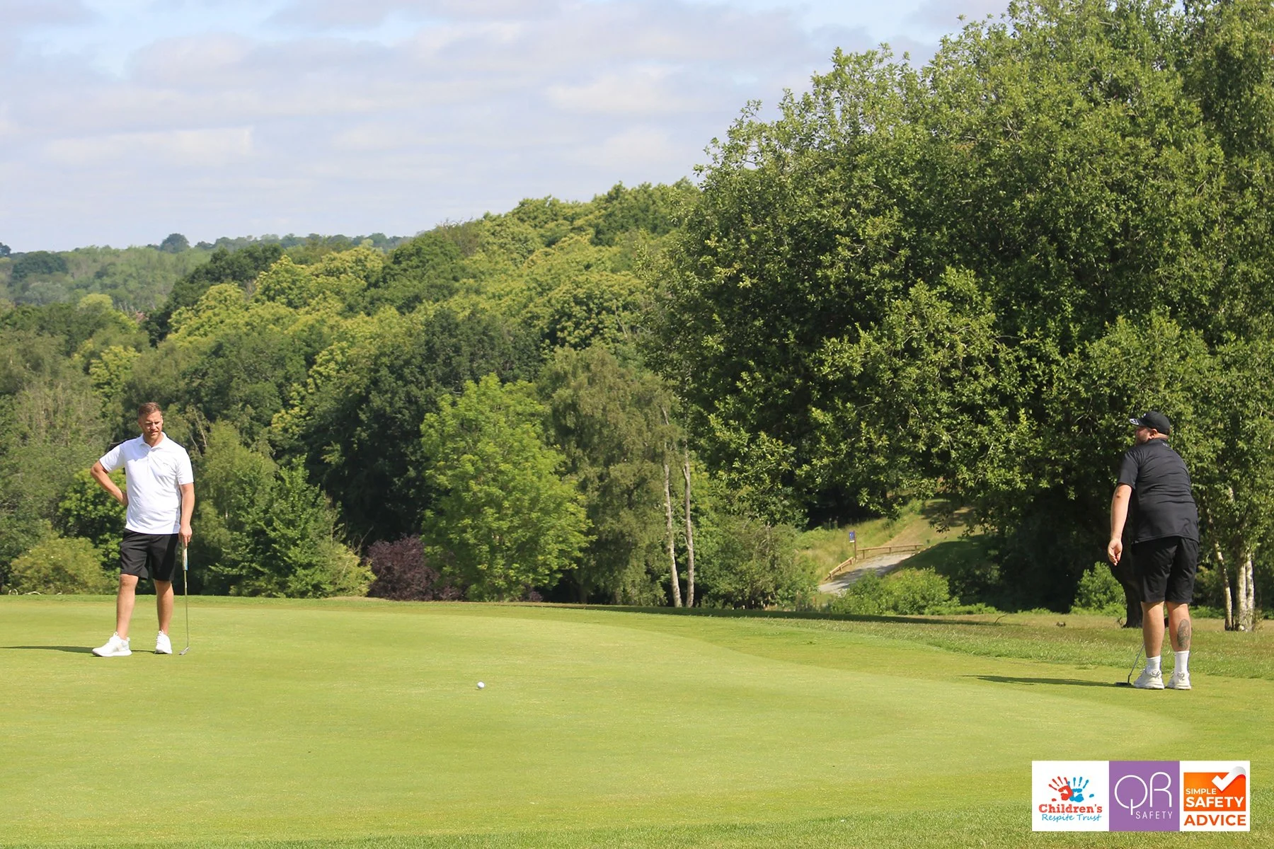 Two men standing on a golf course surrounded by green trees, one preparing to putt as indicated by him holding a golf club close to a golf ball.