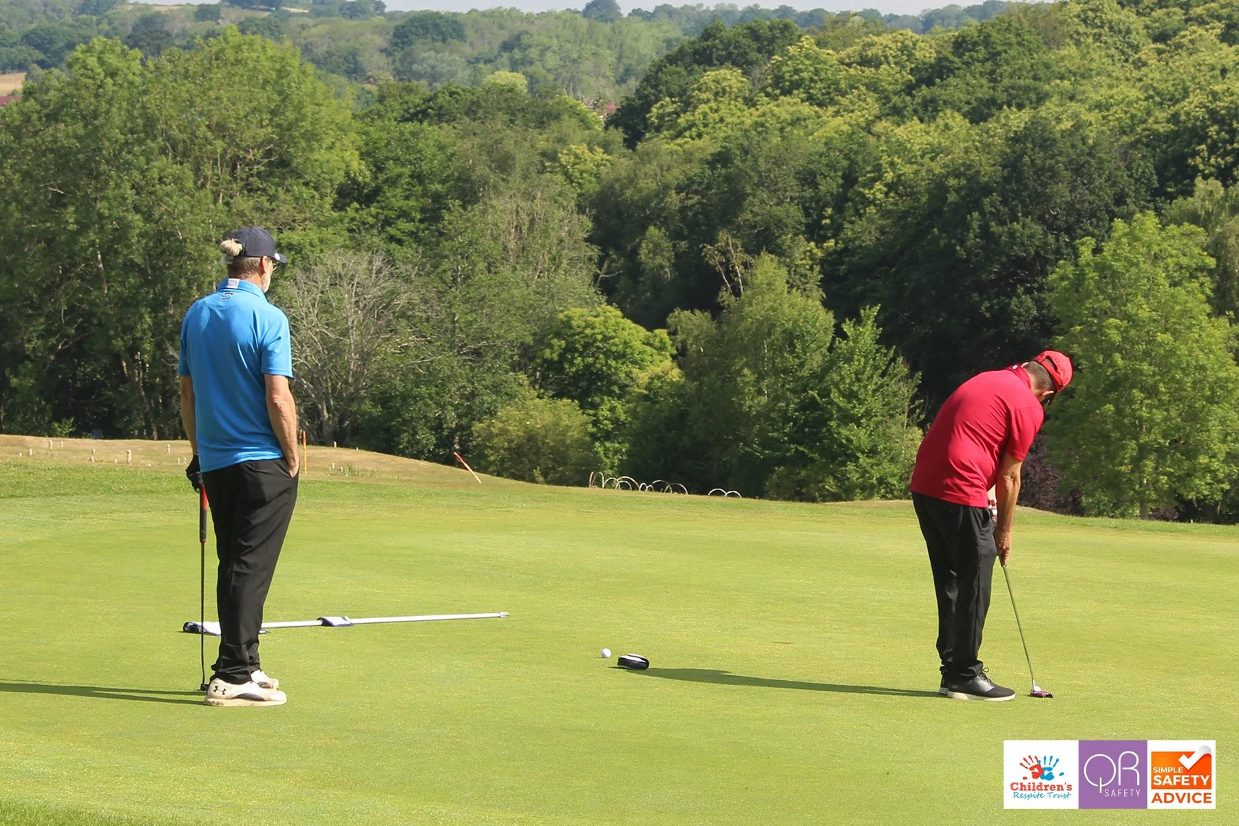Two men playing golf on a green course surrounded by trees, one swinging a golf club, the other watching.