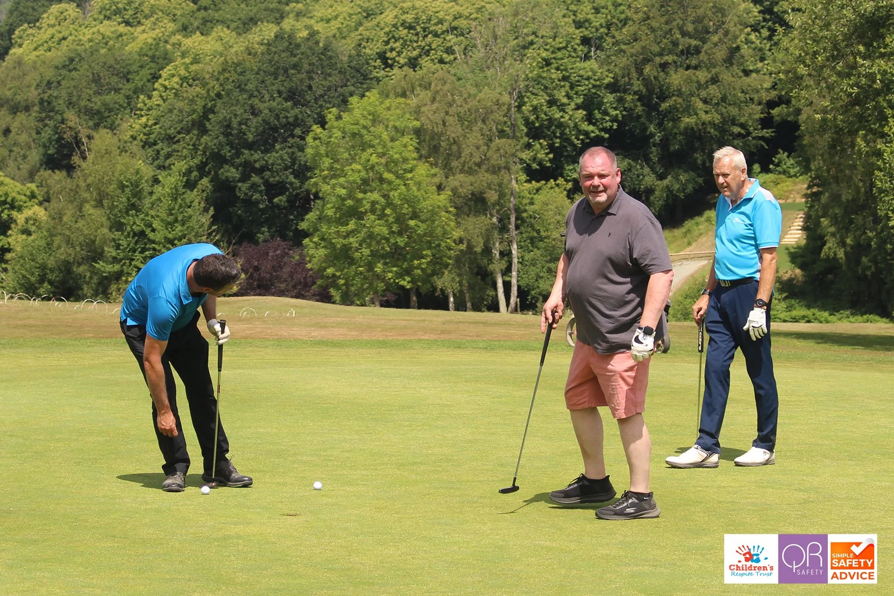 Three men playing golf on a sunny day on a green golf course surrounded by trees, with logos of Children's Response Trust, OP Safety, and Simple Safety Advice in the bottom right corner.