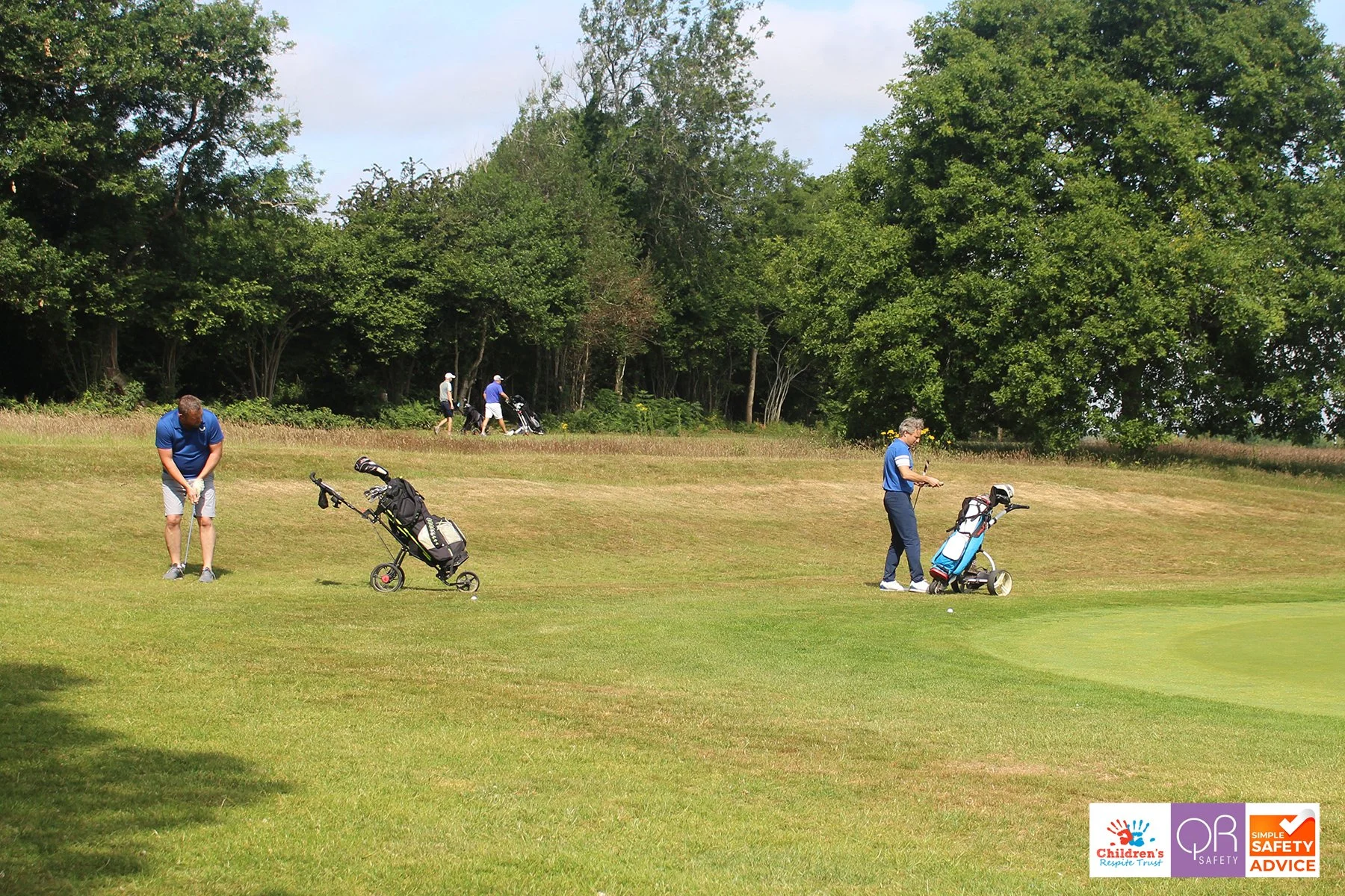 Golfers lining up on a grassy golf course with golf bags on pushcarts, surrounded by trees and a partly cloudy sky.