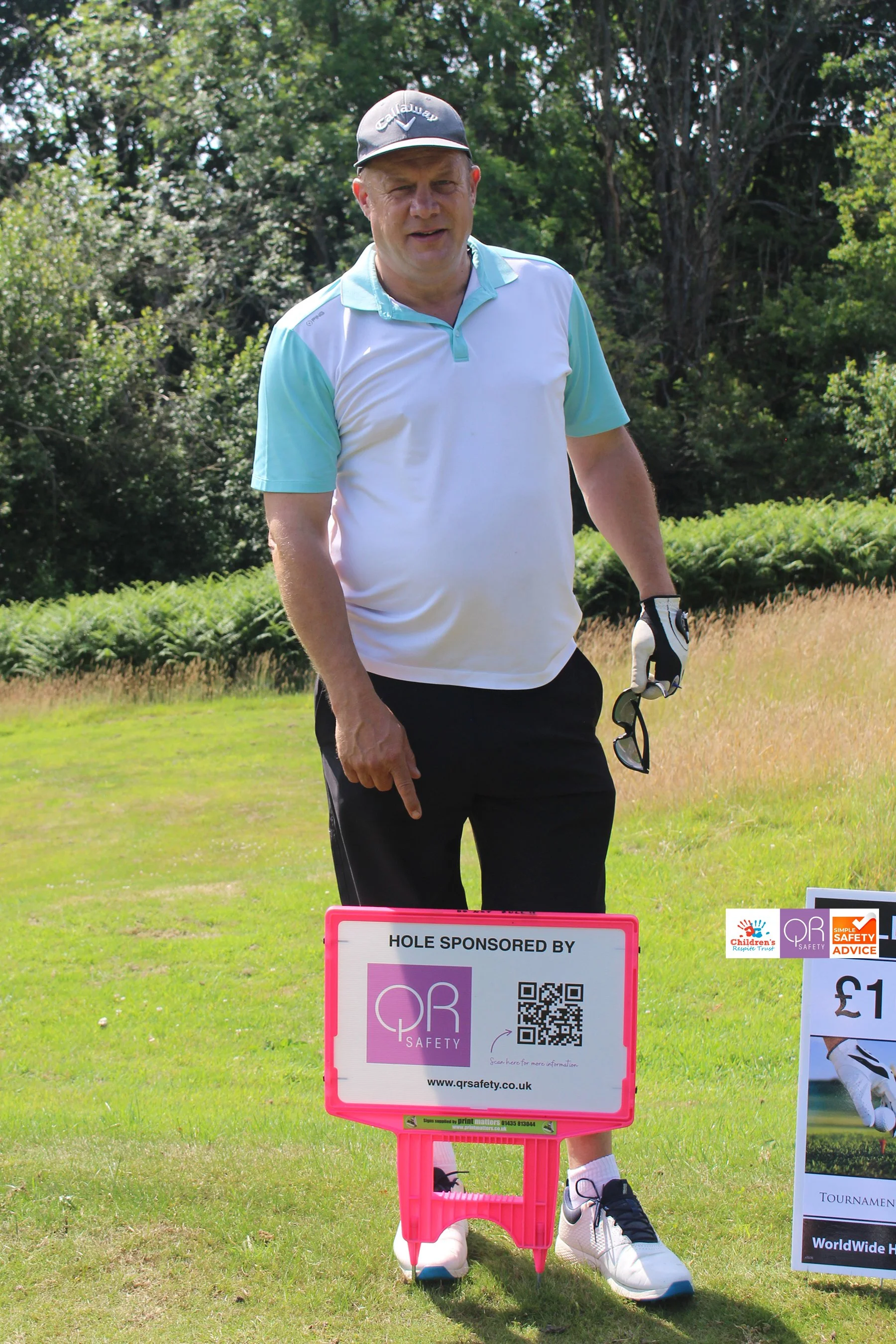 A man on a golf course standing next to a pink sign that reads 'Hole Sponsored by QR Safety' with a QR code and website. The man is wearing a Callaway cap, a white and light blue polo shirt, black pants, and white golf shoes. He is holding golf glass