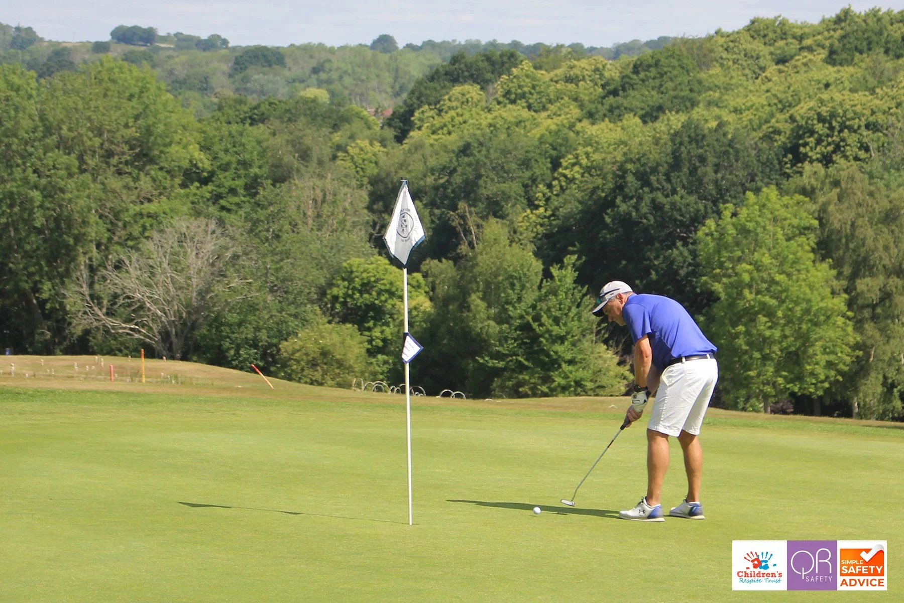A man dressed in a blue shirt, white shorts, and a cap is preparing to putt a golf ball on the green near the hole with a flagstick. The background features lush green trees and a clear, sunny sky.