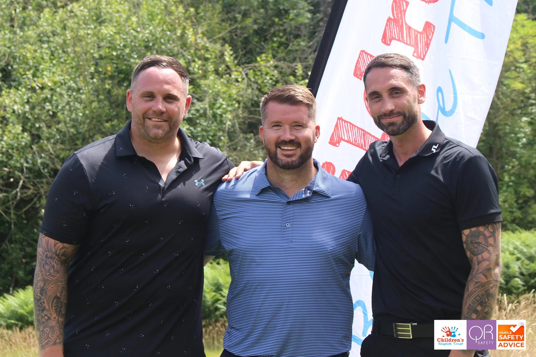 Three men smiling and standing outdoors in front of a white banner with red and blue text. Two are wearing black collared shirts, and one is in a blue striped shirt. They are in a grassy area with trees in the background.