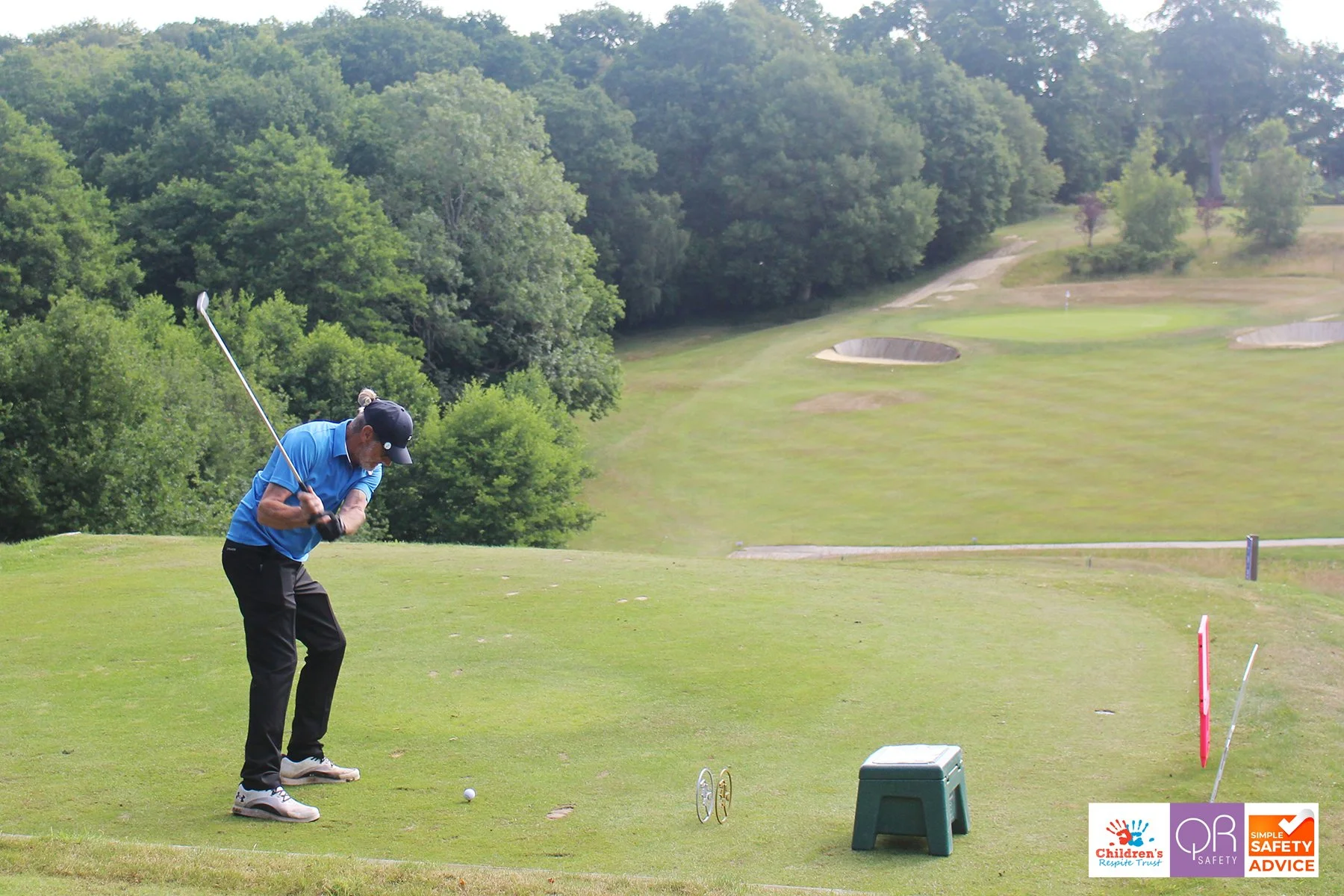 Man in golf attire practicing his swing on a golf course with trees and sand bunkers in the background.