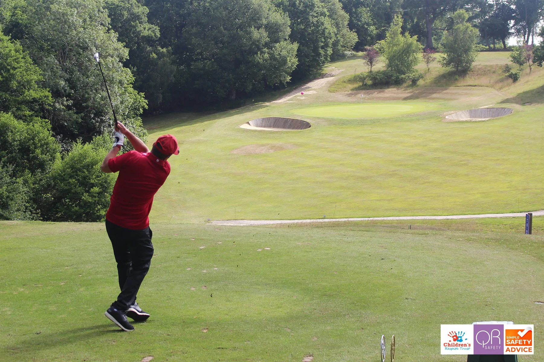 A man in a red shirt and red cap swinging a golf club on a golf course, with trees and sand traps in the background, and safety logos in the bottom right corner.