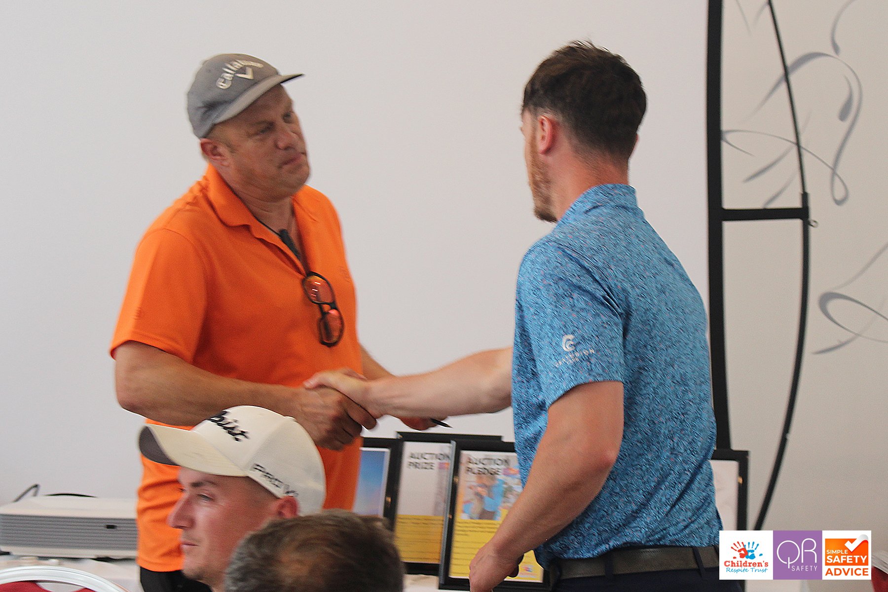 A man in an orange polo shirt and gray cap shaking hands with a young man in a blue shirt, possibly at a formal event or awards ceremony, with certificates or awards on display in the background.