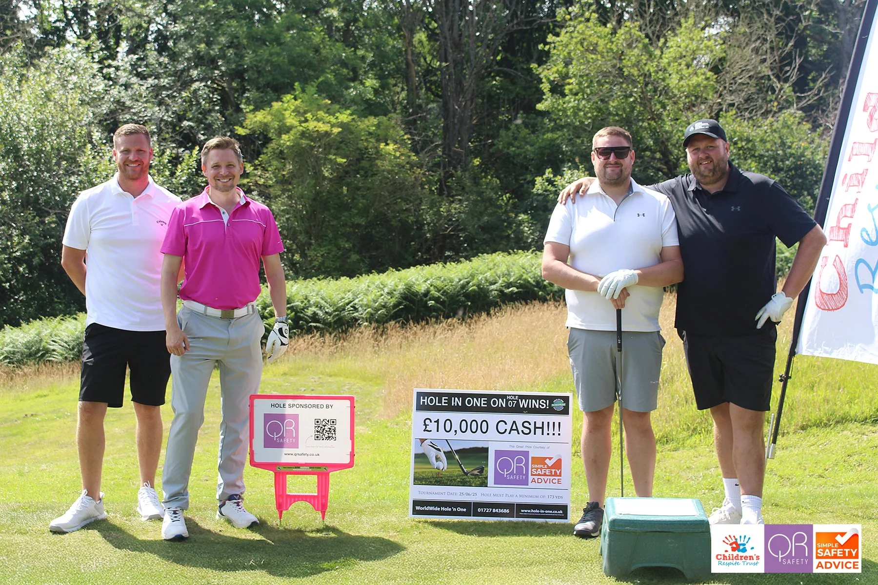 Four men standing on a golf course holding a sign that reads 'Hole in One on 07 wins £10,000 cash!!!' with two of them wearing golf gloves and two smiling at the camera. There is a green box on the ground in front of them, and a banner with safety lo
