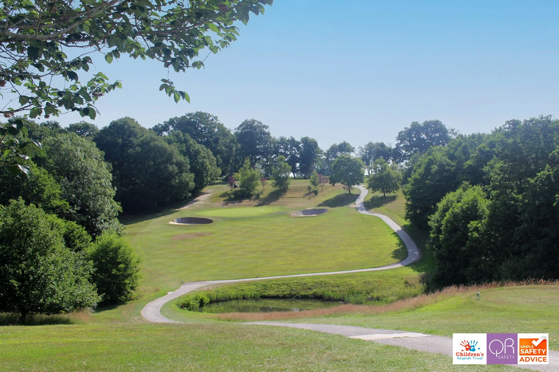 A golf course on a sunny day with trees lining the landscape, winding pathways, a small pond, and two bunkers visible on the grass.