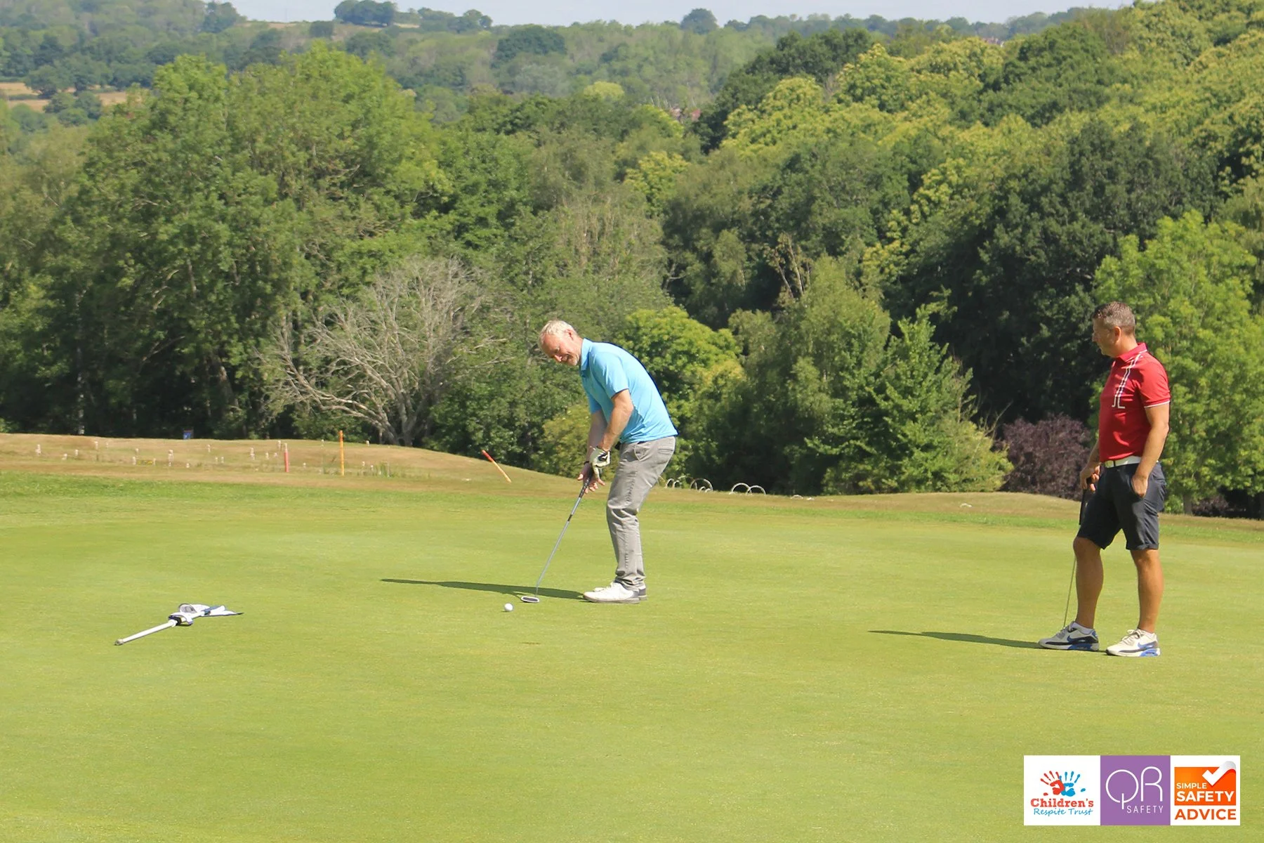 Two men on a golf course, one putting and the other observing, with safety and children's safety logos in the corner.