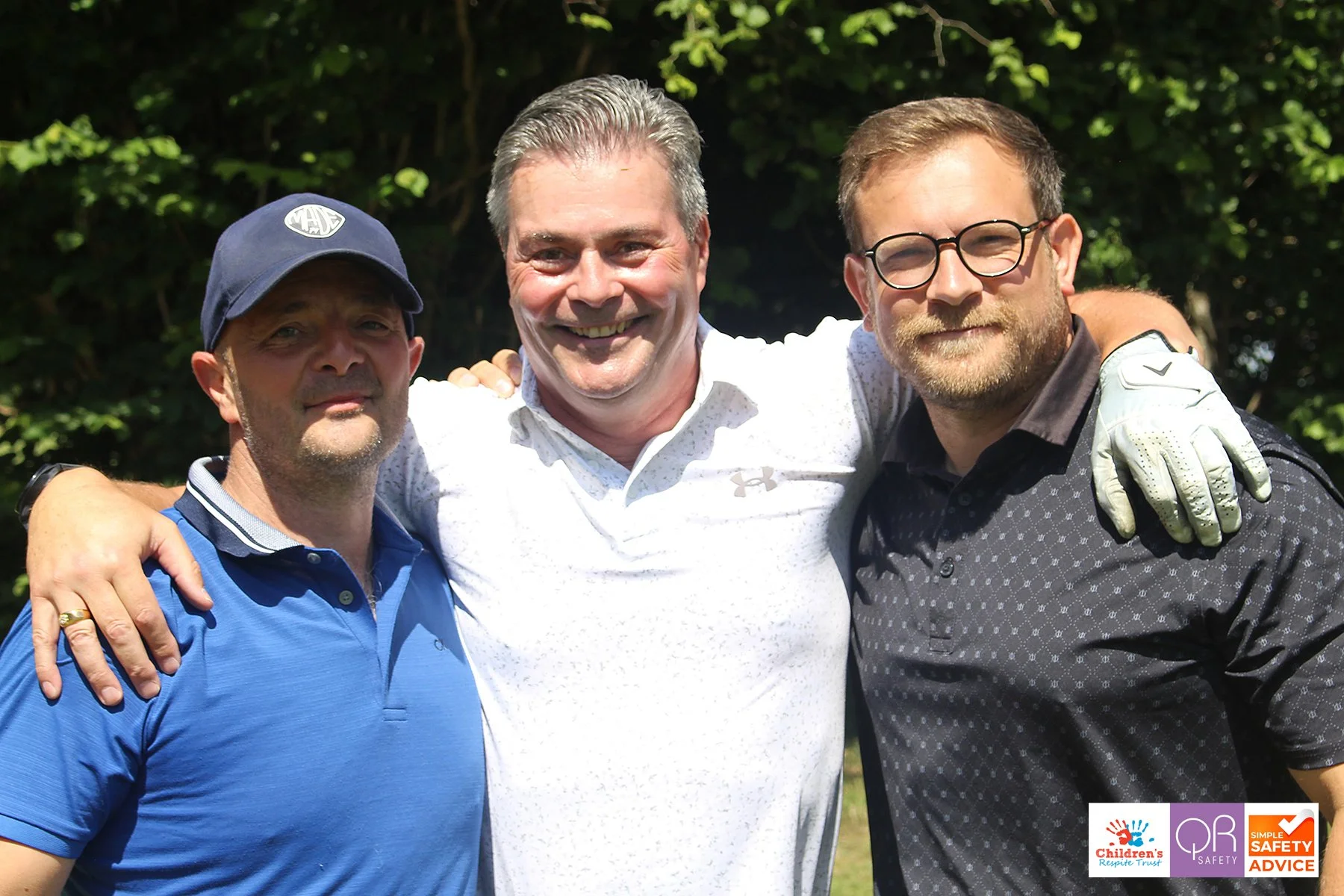 Three men standing outdoors with green trees in the background, smiling and putting their arms around each other, dressed casually, one wearing a baseball cap, one with glasses, and the other smiling broadly.