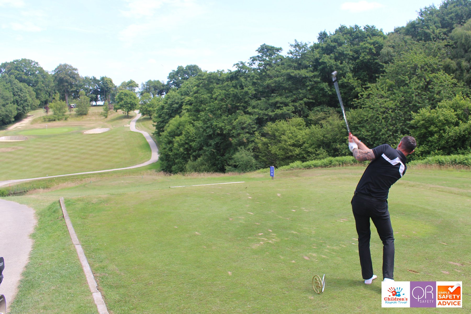 A man with a tattooed right arm taking a powerful golf swing on a lush, green golf course with trees and rolling hills in the background.
