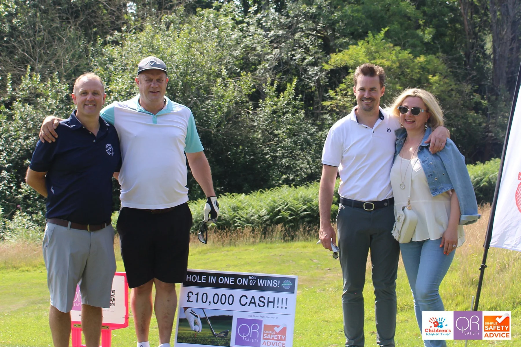 Four people standing outdoors on a golf course, celebrating a win with a sign that reads '£10,000 Cash!!!' and logos for children's respite trust and safety advice.