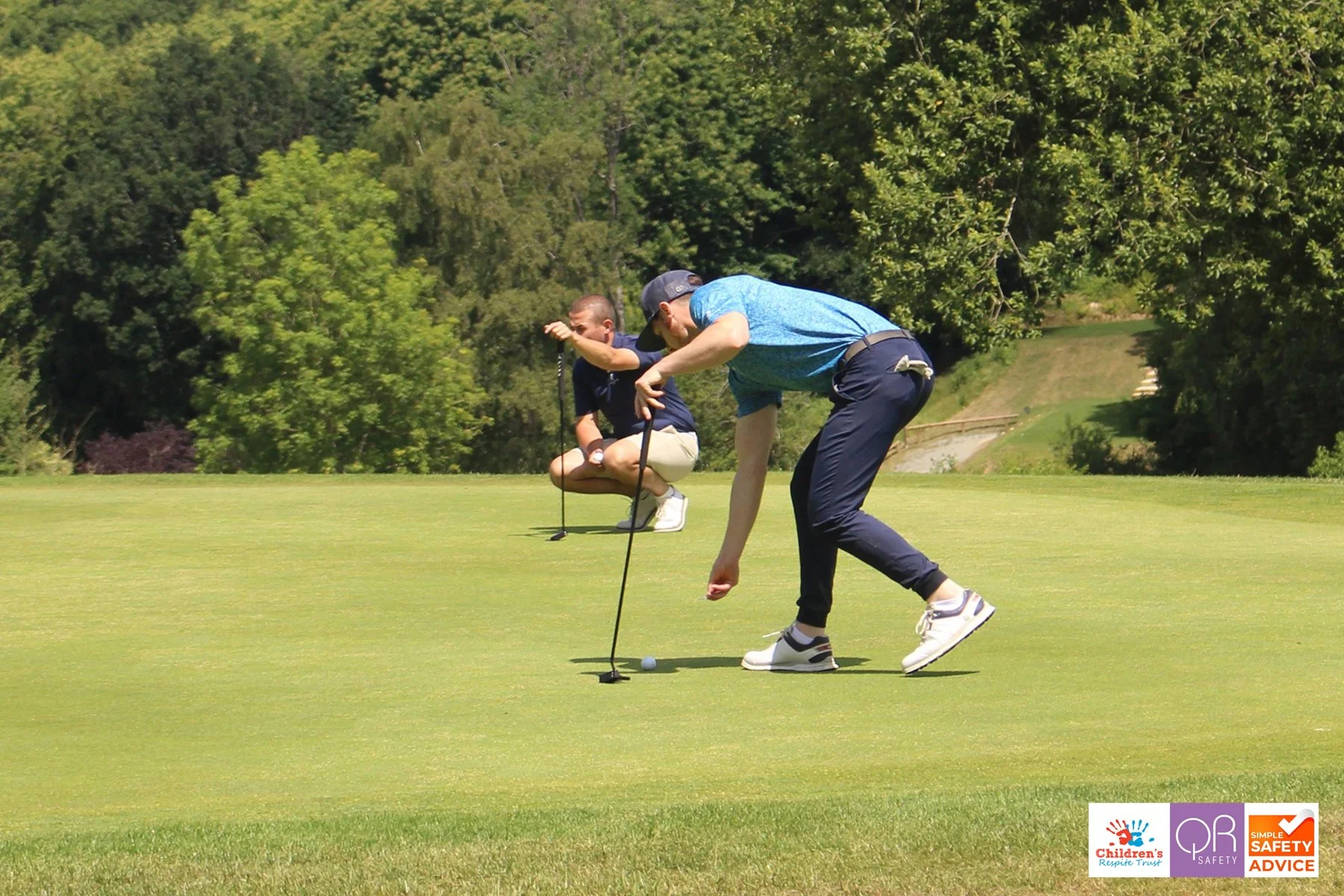 Two men playing golf on a bright, sunny day, one about to putt a golf ball on the green with a golf club, and the other squatting behind him. Trees and a hill in the background.