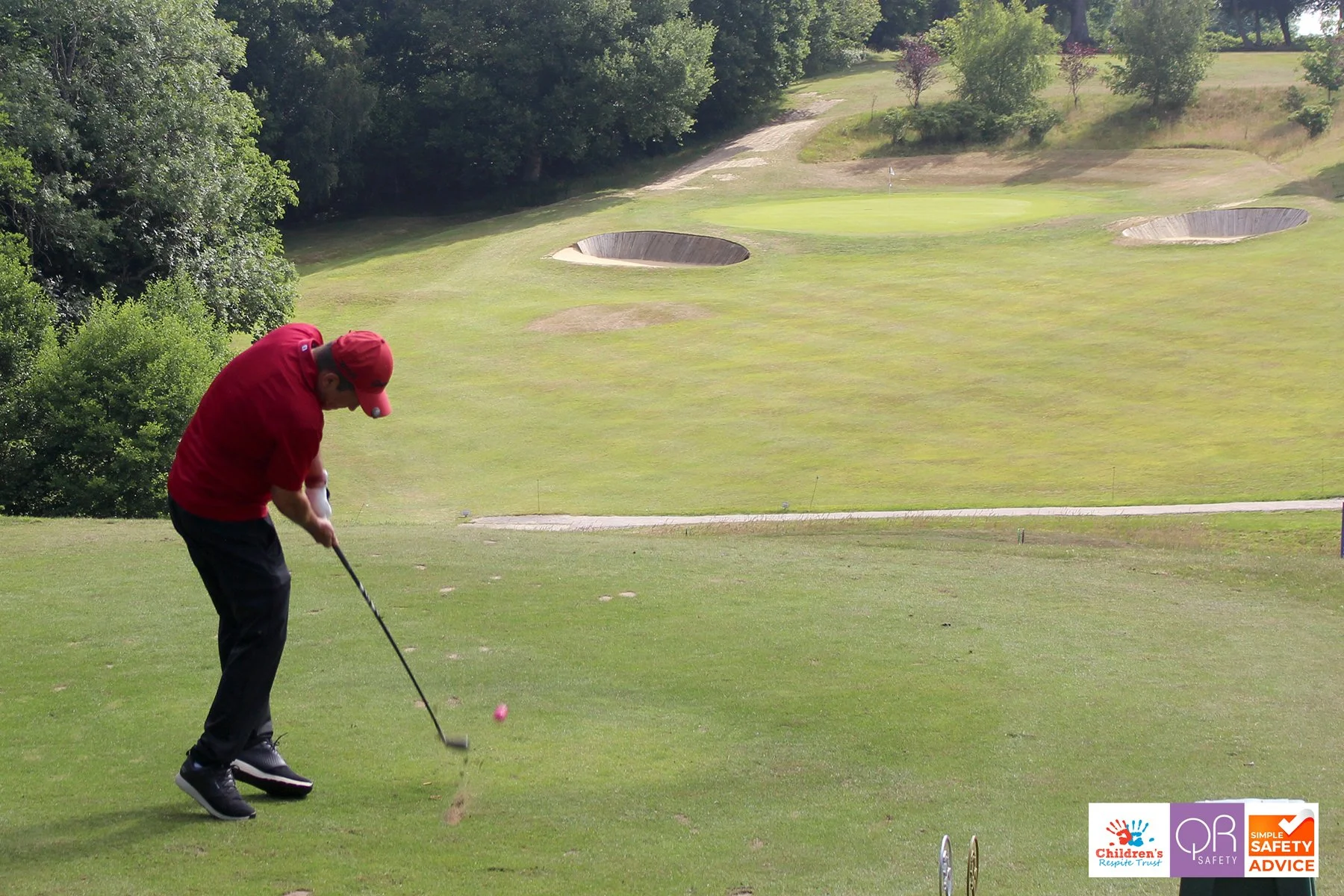 A man in a red shirt and red cap prepares to hit a golf ball on a golf course with trees and sand bunkers in the background.