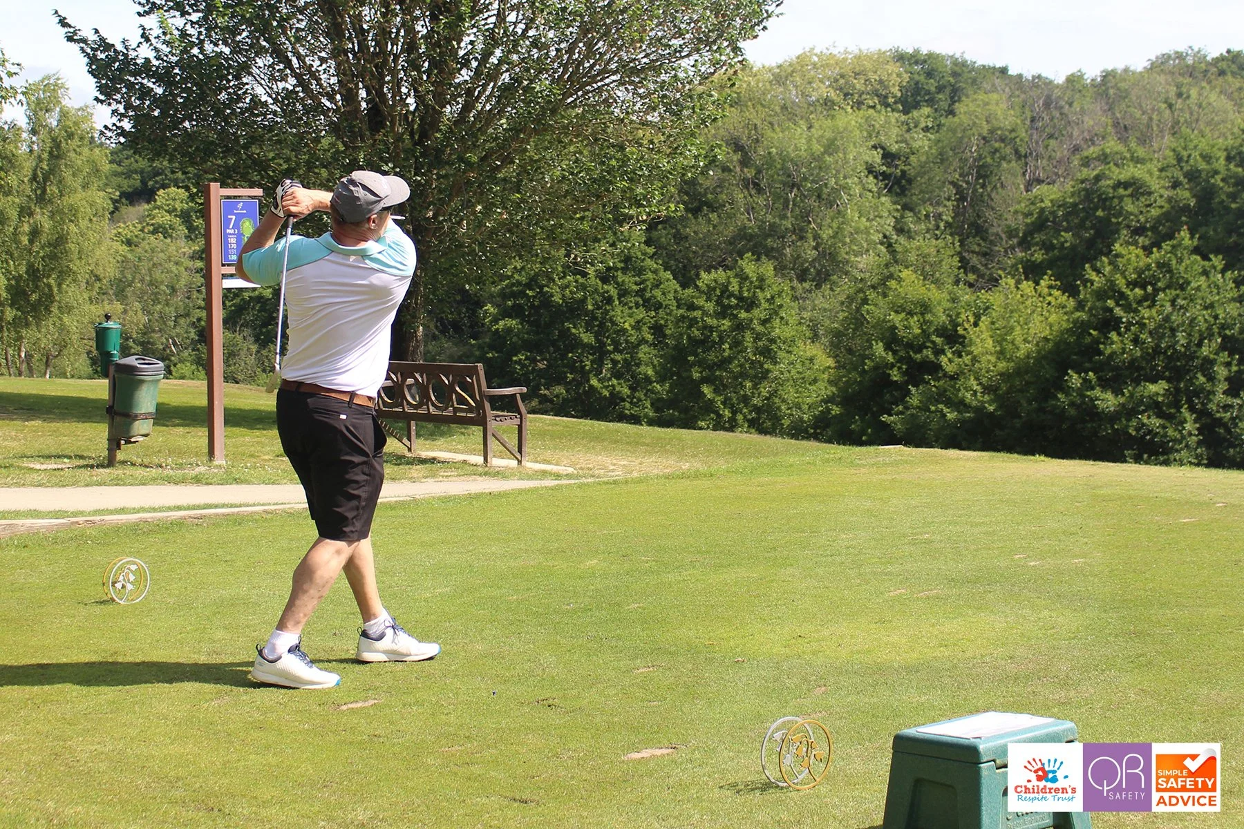 A man in a white and teal shirt, black shorts, and a gray cap swings a golf club on a golf course with green grass and trees in the background.