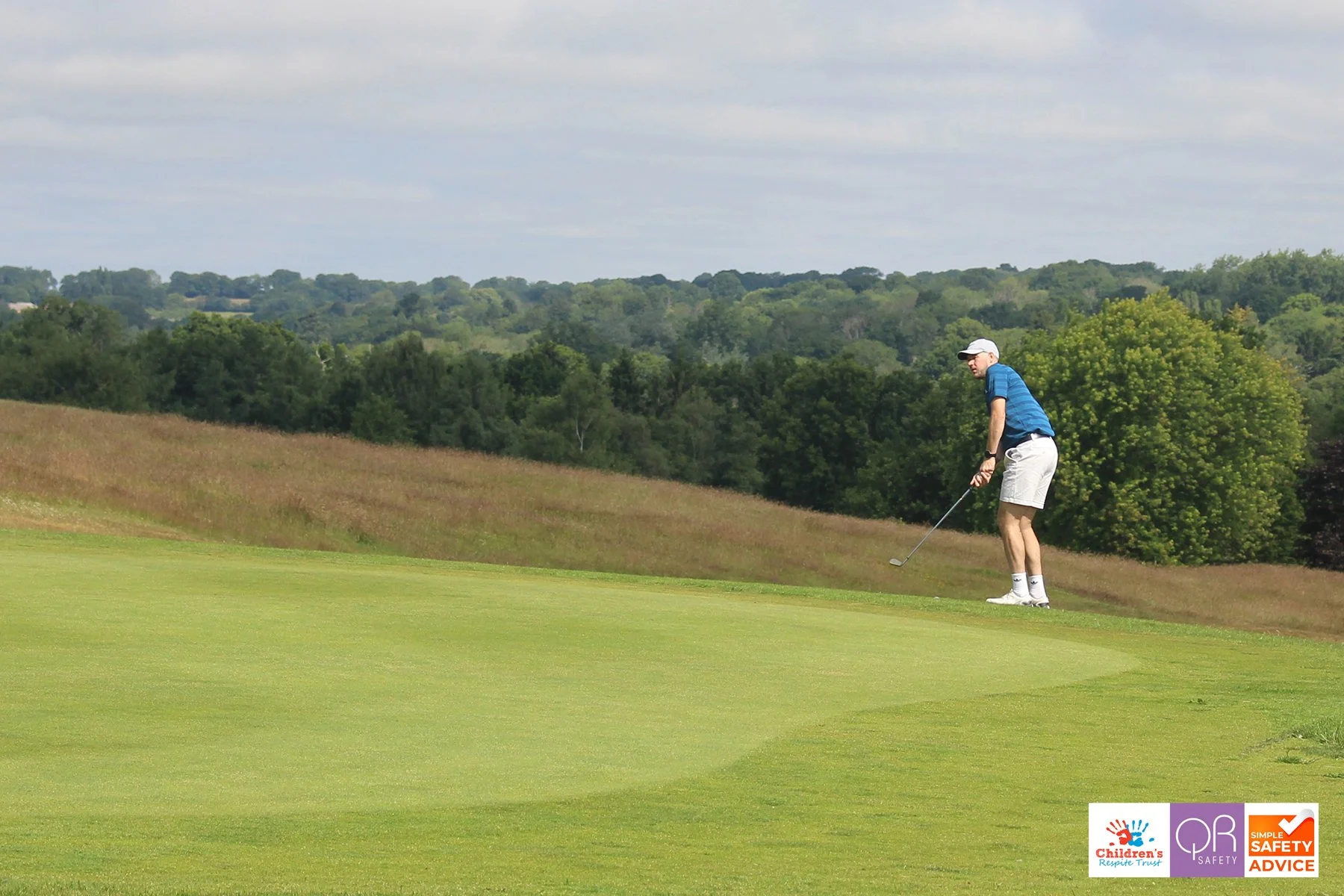 A man in a blue shirt, khaki shorts, white cap, and white shoes playing golf on a green course with trees and hills in the background.