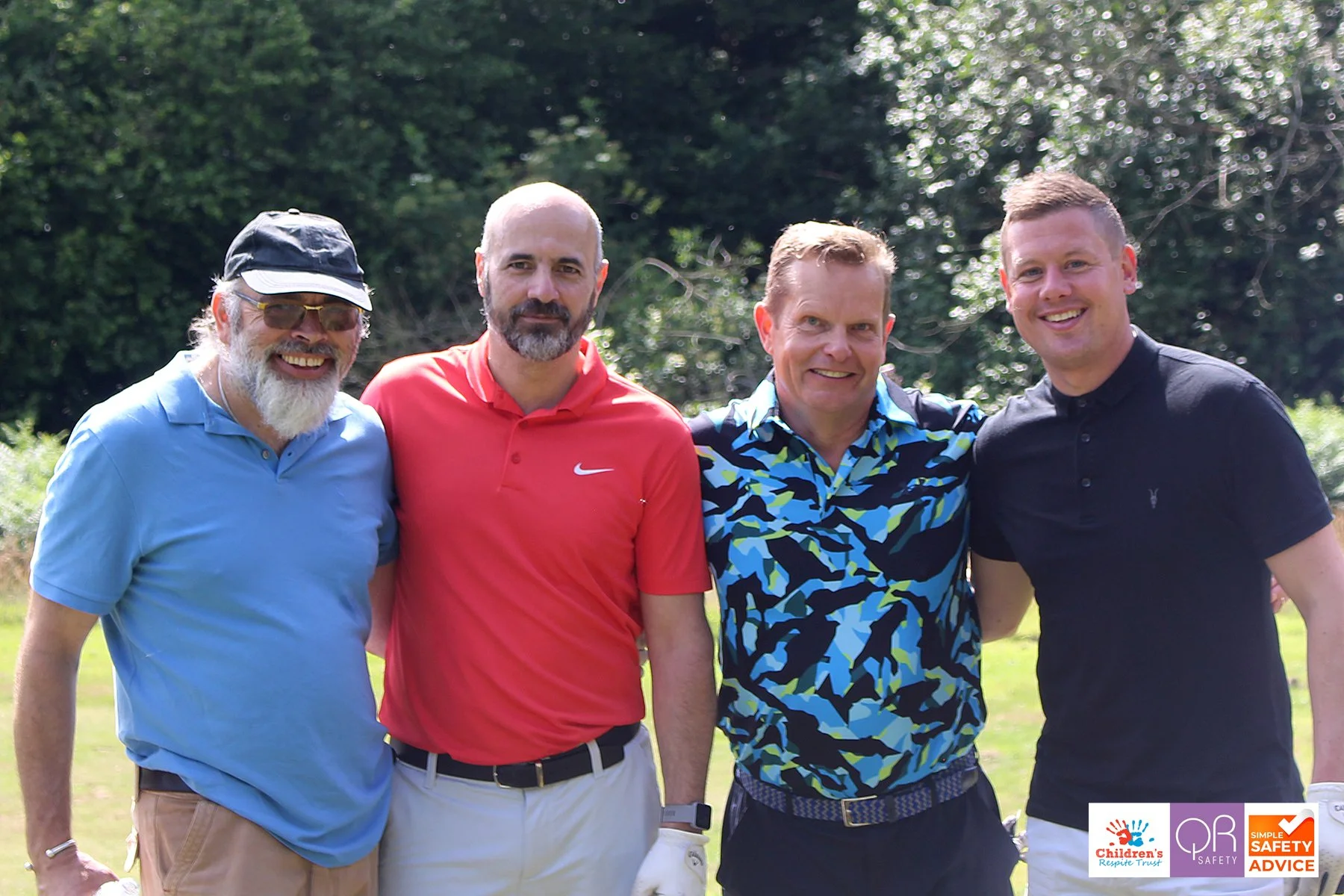 Four men standing outdoors on a golf course, smiling with their arms around each other's shoulders, with trees in the background.