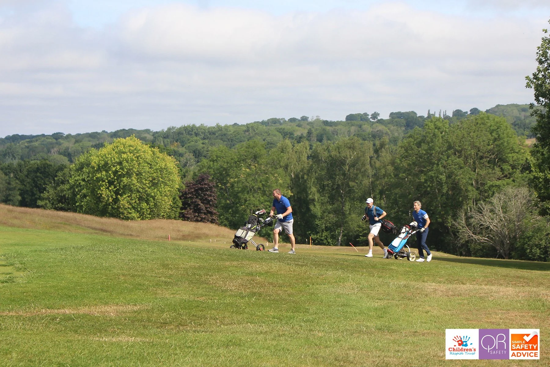 Three men walking on a golf course with golf carts, surrounded by trees and hills, on a partly cloudy day.