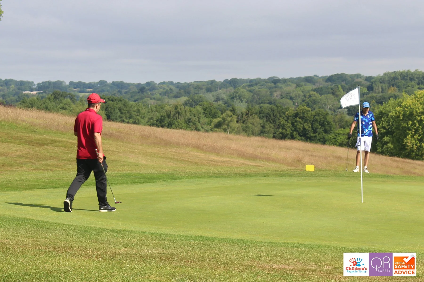 Two men, one in a red shirt and black pants and the other in a blue Hawaiian shirt and white shorts, are on a golf course. One is putting while the other is standing nearby. A white flag with a logo is on the green, and forested hills are in the back