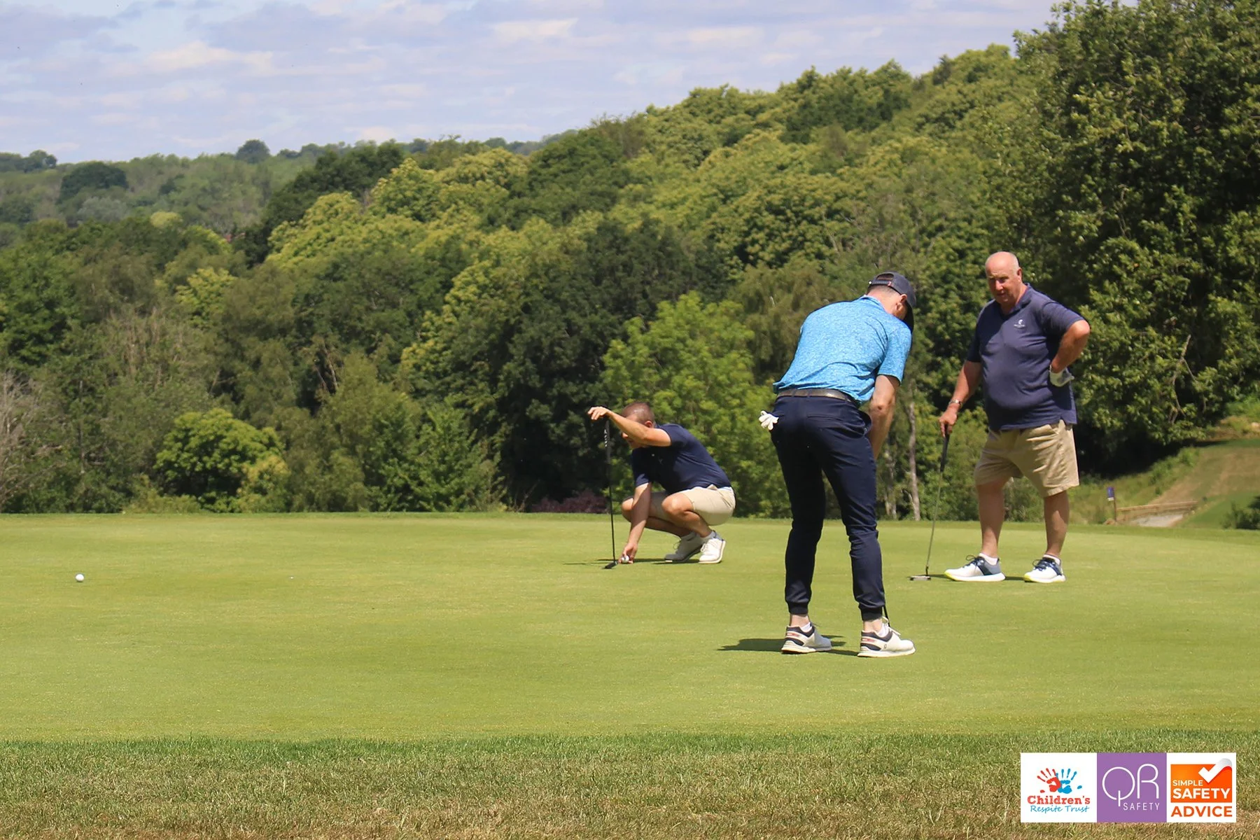 Three men playing golf on a green golf course with a lush, green, tree-covered landscape and blue sky in the background.
