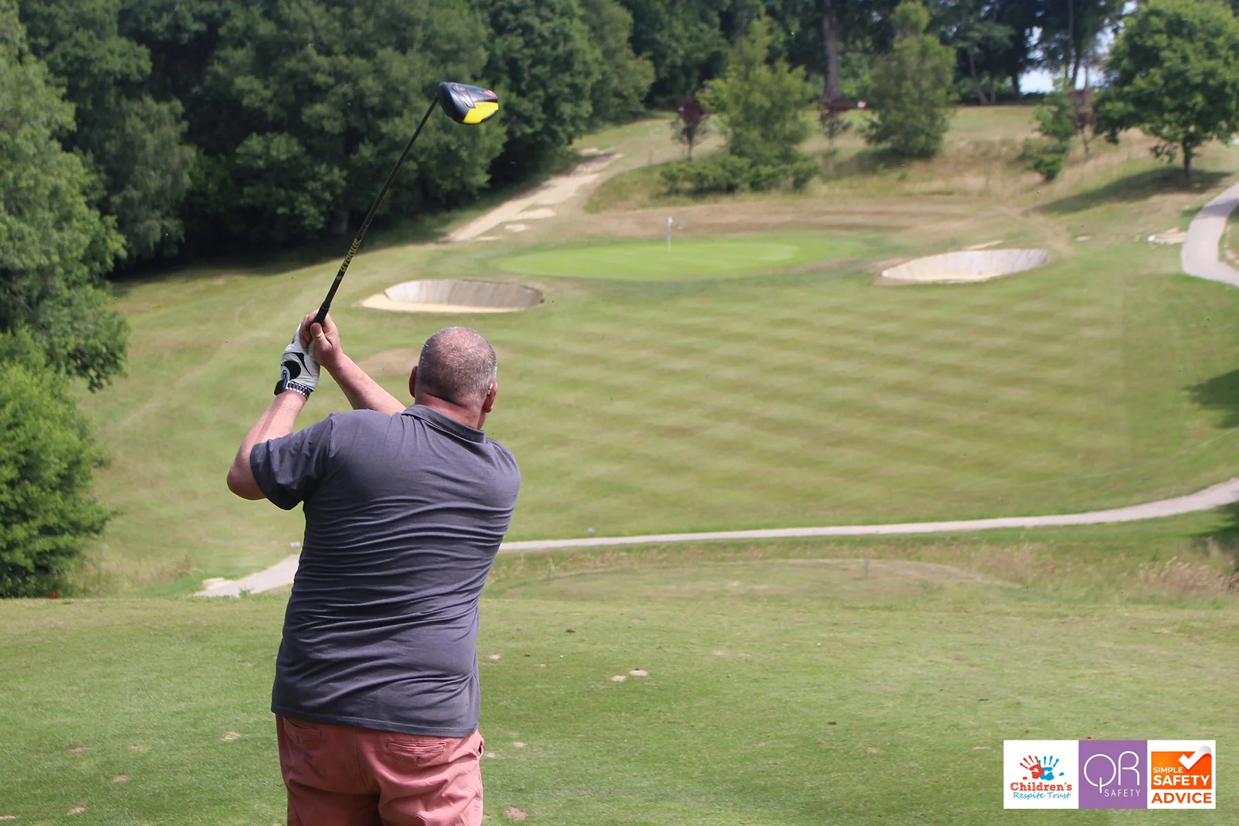 A man with a gray shirt and pink shorts swinging a golf club on a golf course with green trees and bunkers in the background