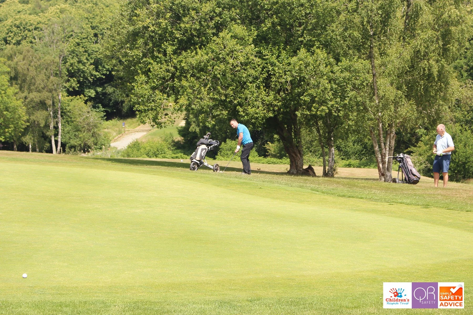 Two men on a golf course, one preparing to hit a golf ball, with golf bags and trees in the background.